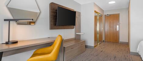Una habitación de hotel con escritorio, una silla amarilla, televisor montado y una puerta de madera.