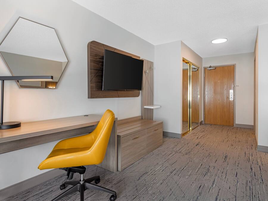 A hotel room with a desk, a yellow chair, a mounted TV, and a wooden door.