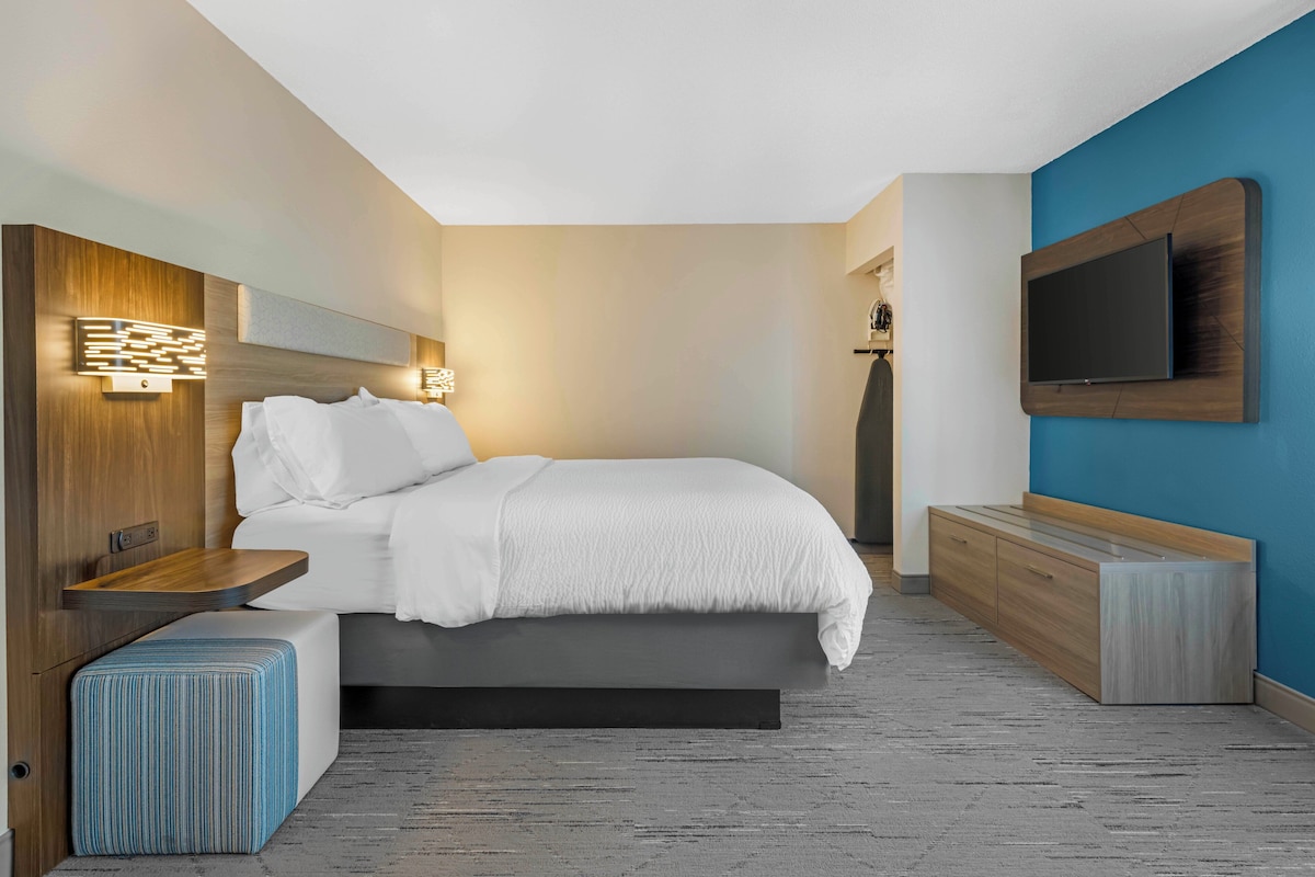 A modern hotel room with a bed, bedside table, bench, TV, and wall-mounted lighting.