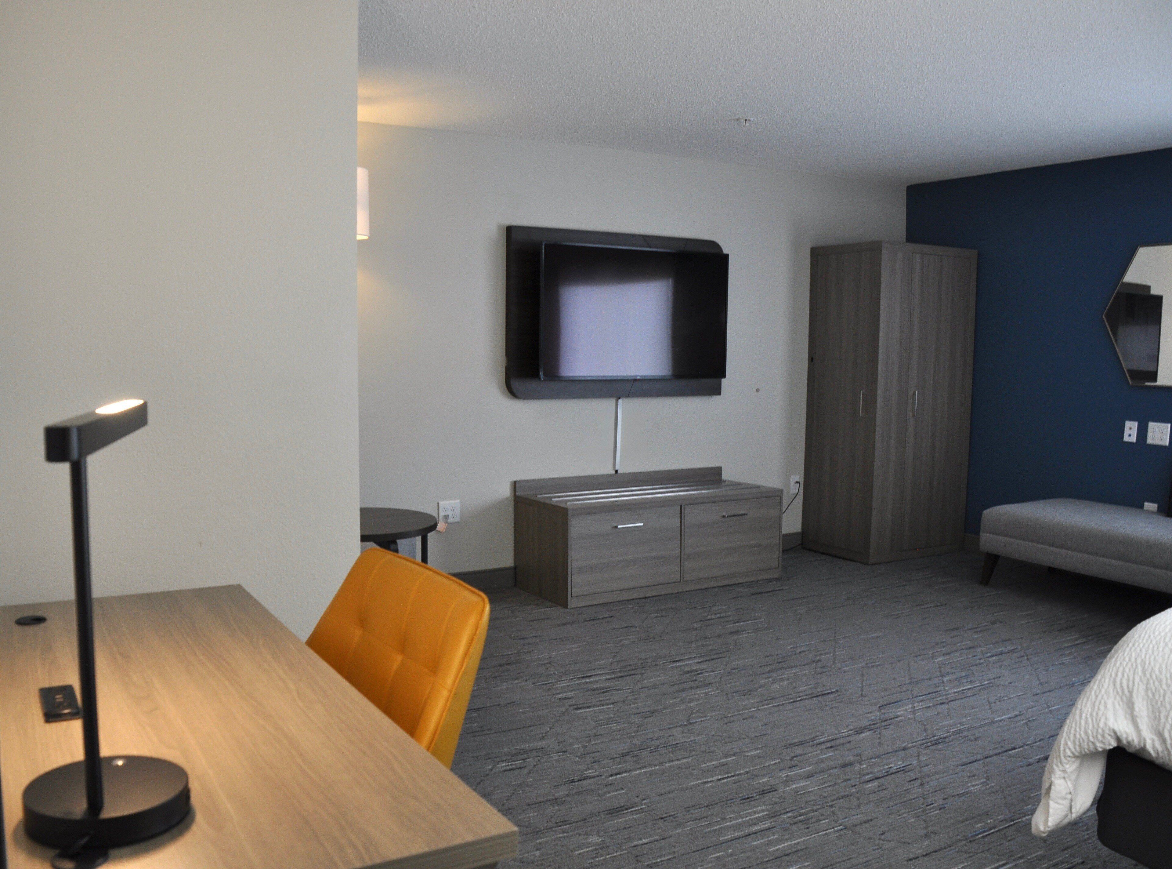 Una habitación de hotel moderna con una televisión de pantalla plana montada en la pared, un escritorio de madera con una lámpara, un sofá y un armario.
