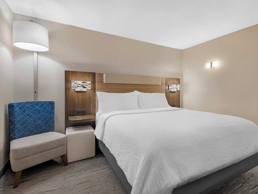 A hotel room with a bed, a chair, a nightstand, a lamp, and a wall-mounted light fixture.