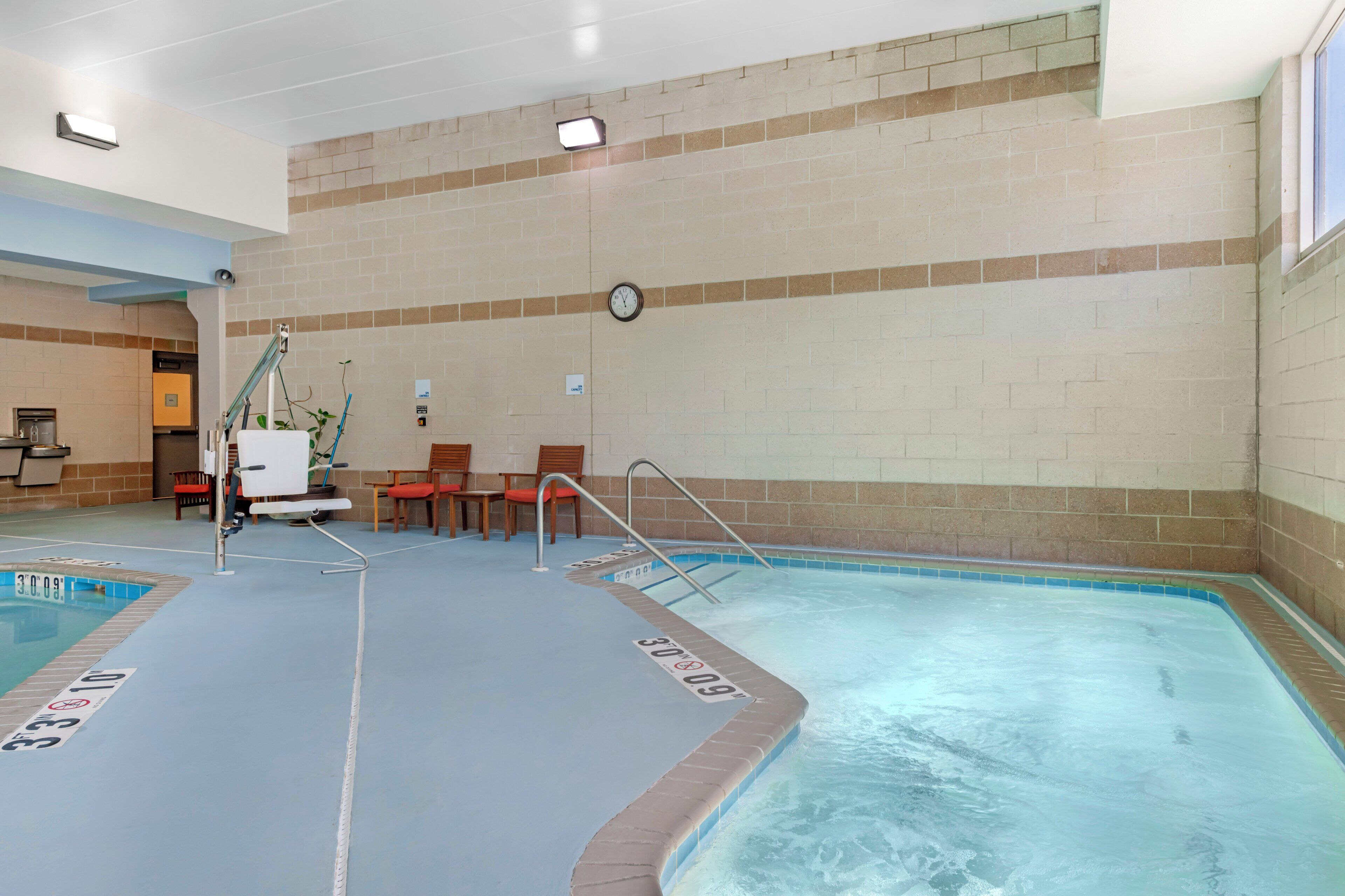 An indoor swimming pool with a clock on the wall, a lifeguard stand, and a seating area with chairs.