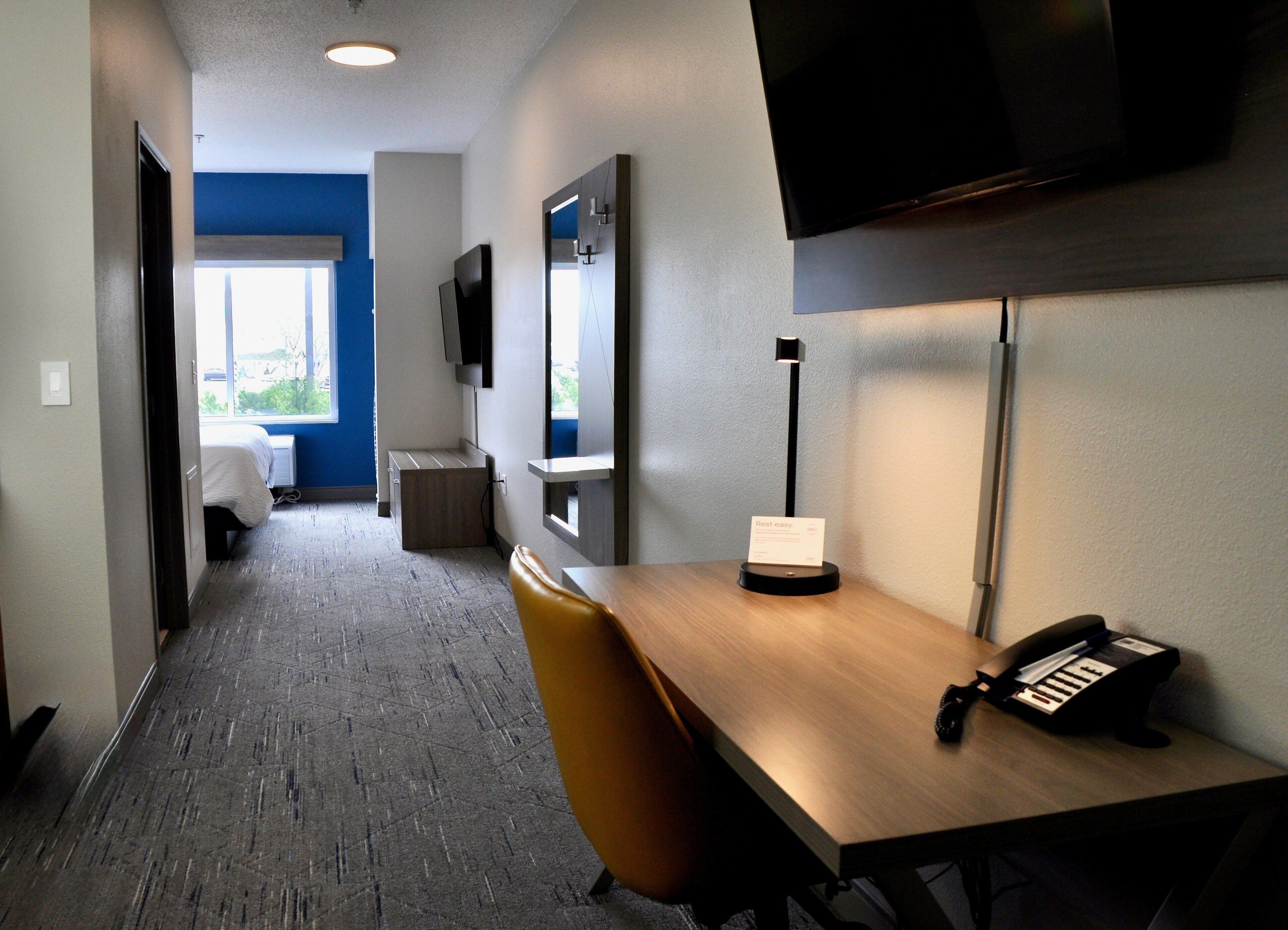 Une chambre d’hôtel avec un bureau en bois, un téléphone, une lampe, une chaise, un téléviseur fixé au mur et un lit en arrière-plan.
