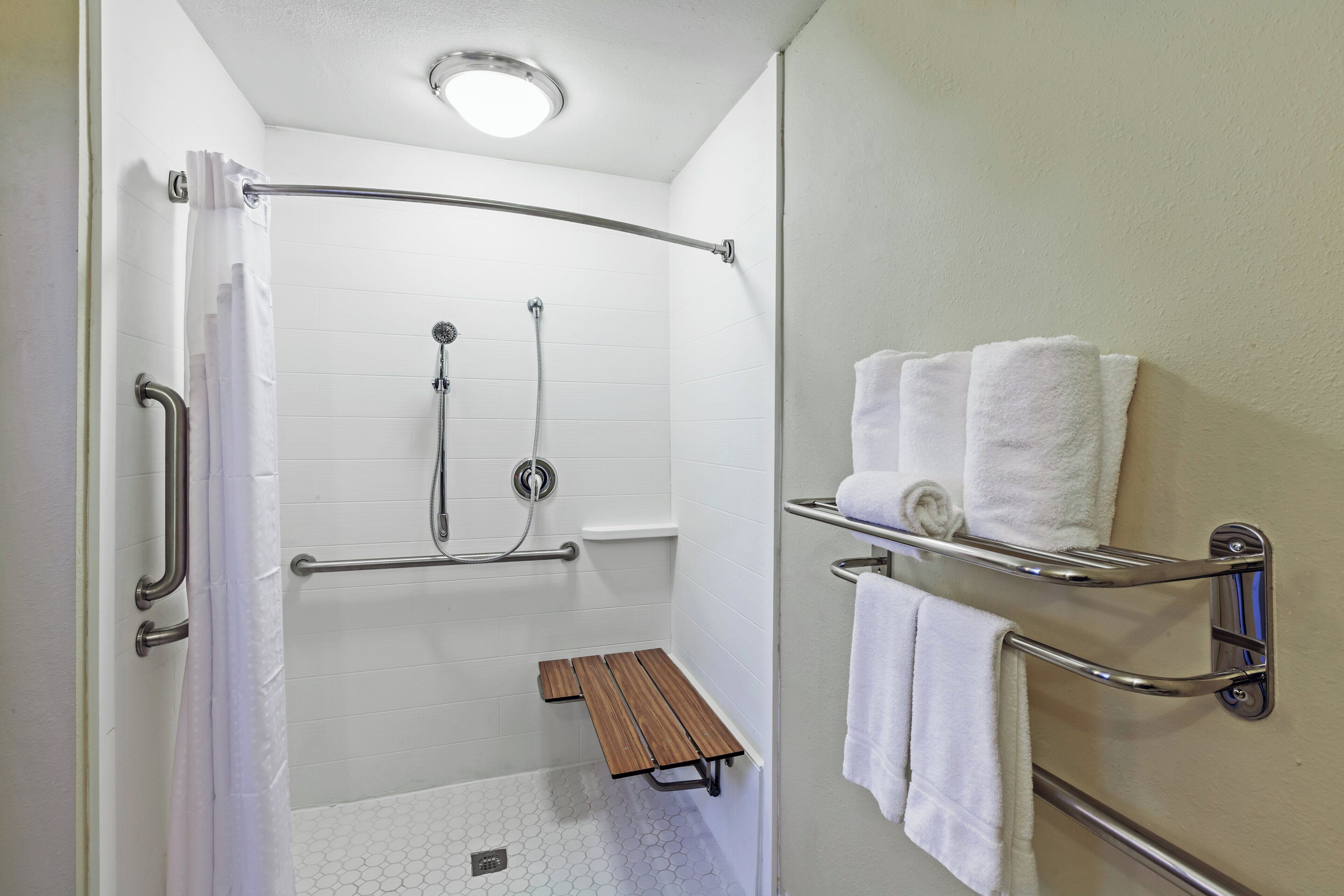A bathroom with a shower, grab bars, a bench, and a towel rack.