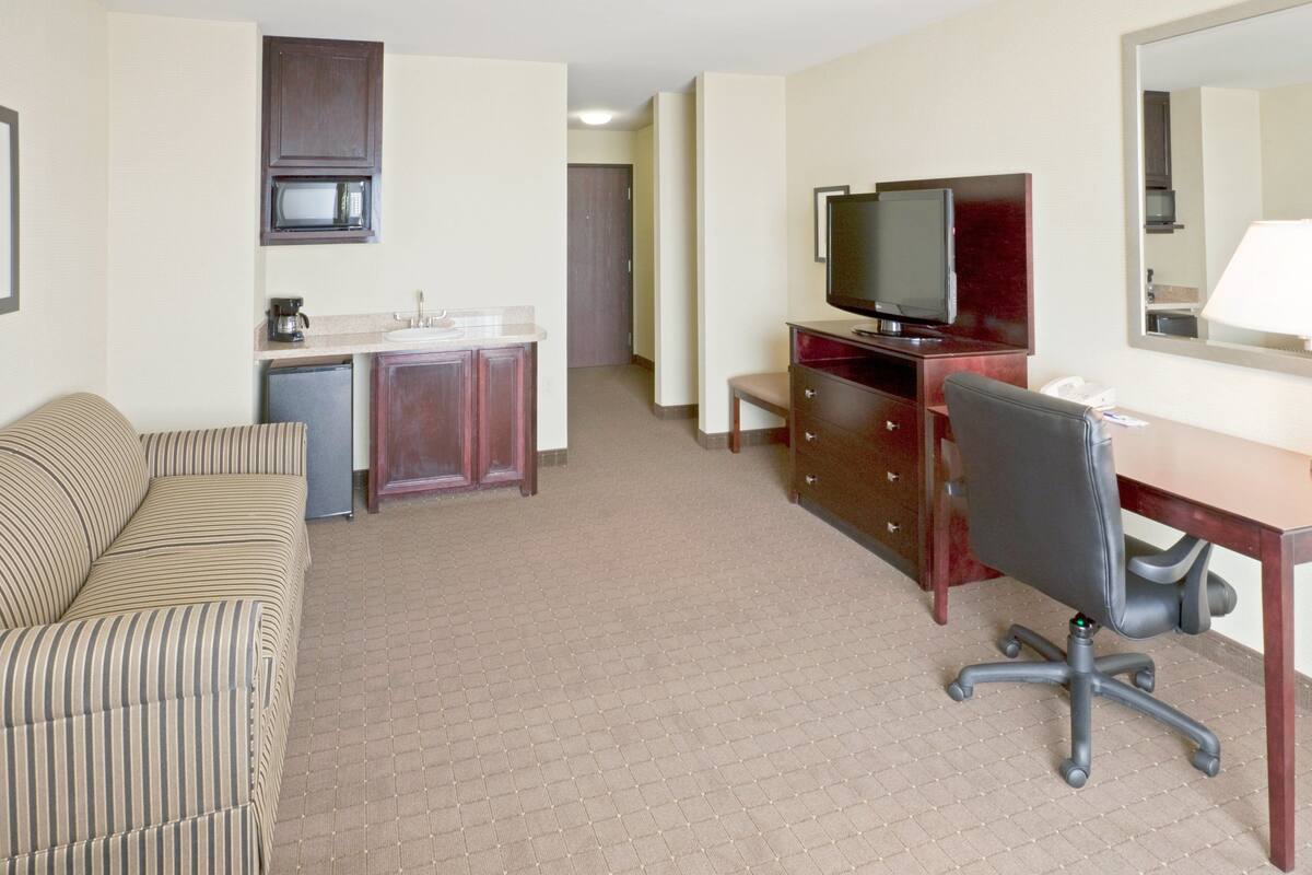 A hotel room with a sofa, a desk with a chair, a television, a mirror, and a kitchenette with a microwave.