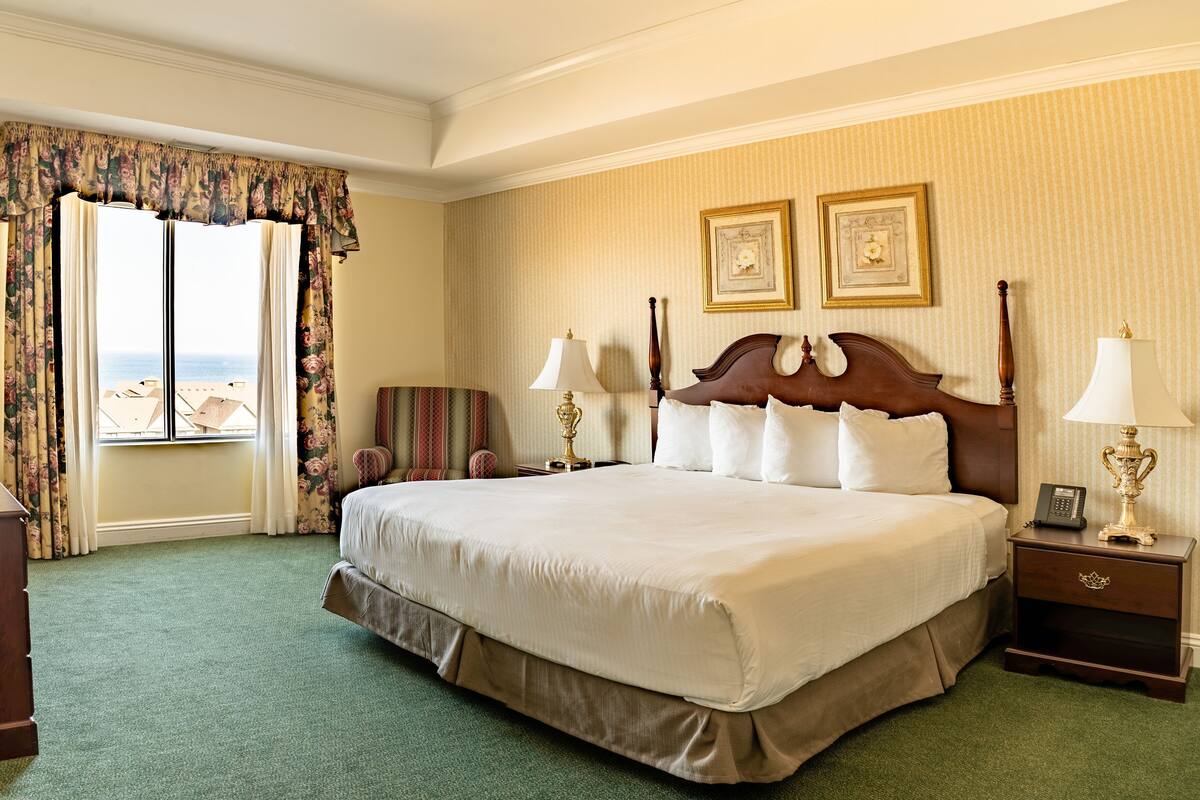 A hotel room with a large bed, two bedside lamps, a nightstand, a chair, a desk with a telephone, and a view of the sea through the window.