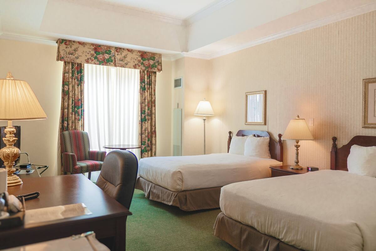 A hotel room with two beds, a desk, a chair, and a lamp.