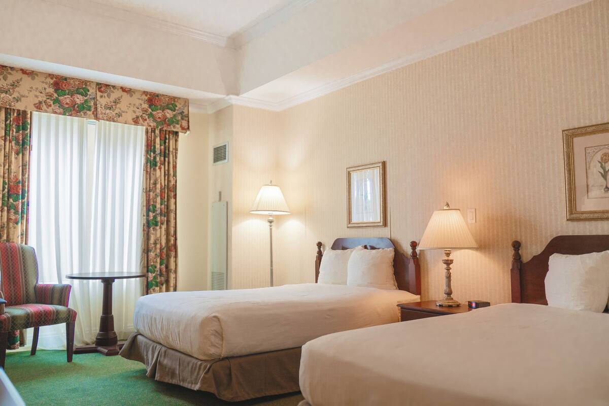 A hotel room with two beds, a chair, a table, lamps, and curtains.
