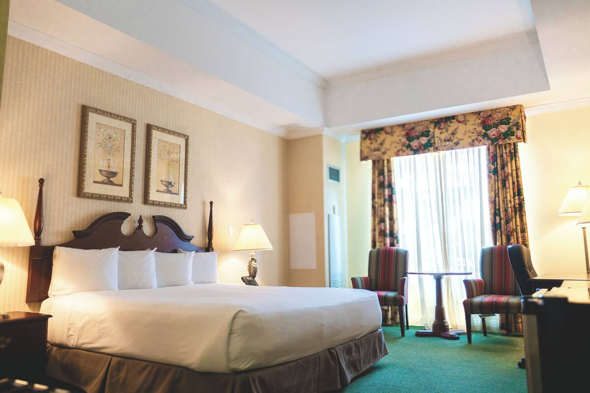 A hotel room with a large bed, two bedside tables with lamps, a small table with chairs, and a window with curtains.