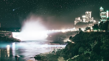 Nachtsicht auf eine Stadt mit einem Wasserfall, beleuchteten Gebäuden und einem Sternenhimmel.