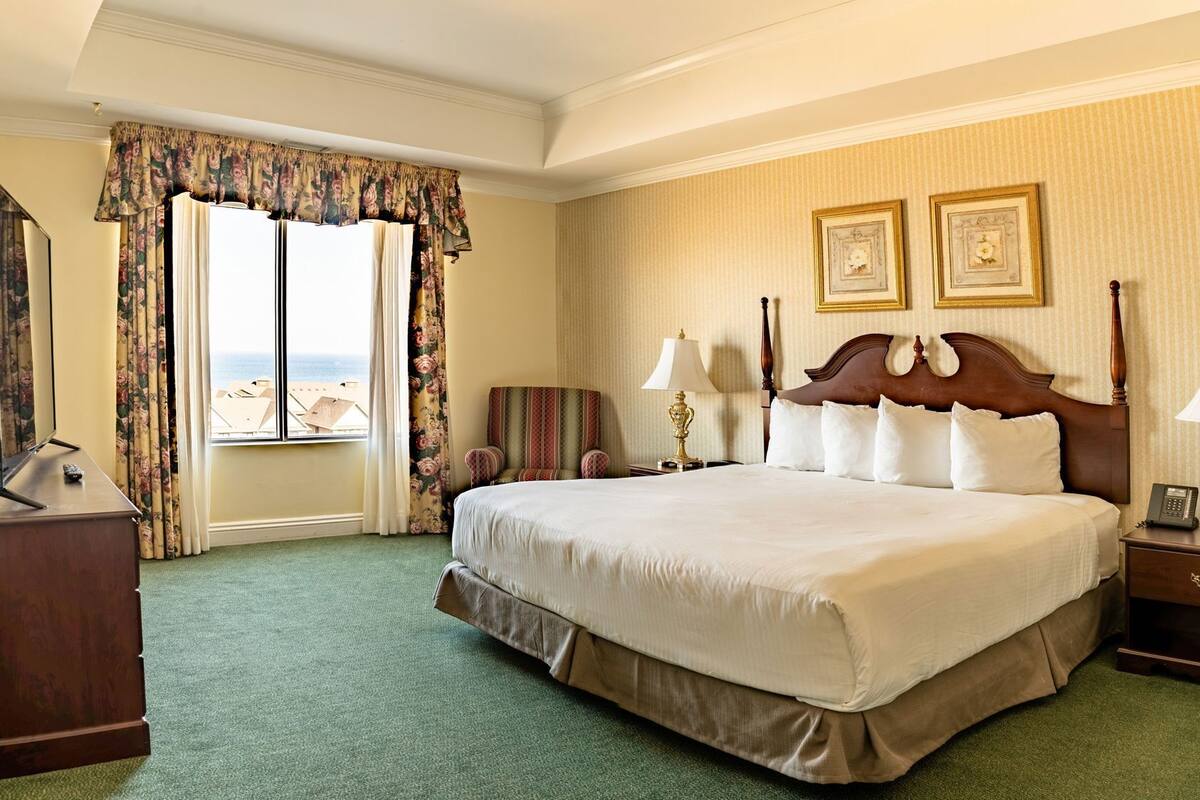 A hotel room with a large bed, two bedside lamps, a telephone, a chair, and a view of the sea.