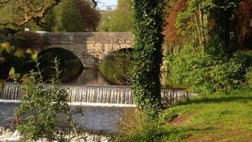 A stone bridge over a river with a small waterfall, surrounded by greenery and trees.