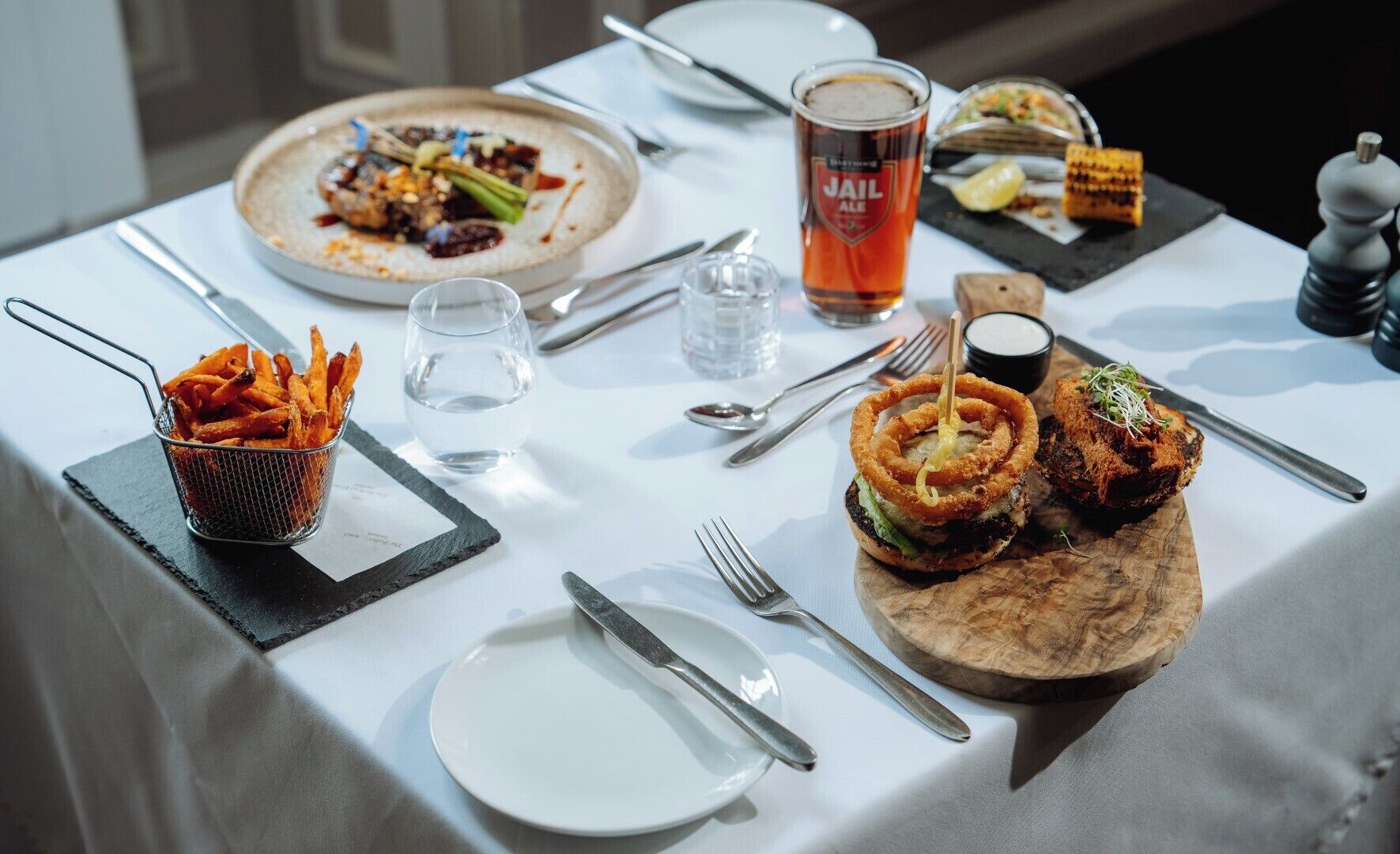 A dining table with a variety of dishes, including a burger with onion rings, a plate of fries, and a glass of beer.