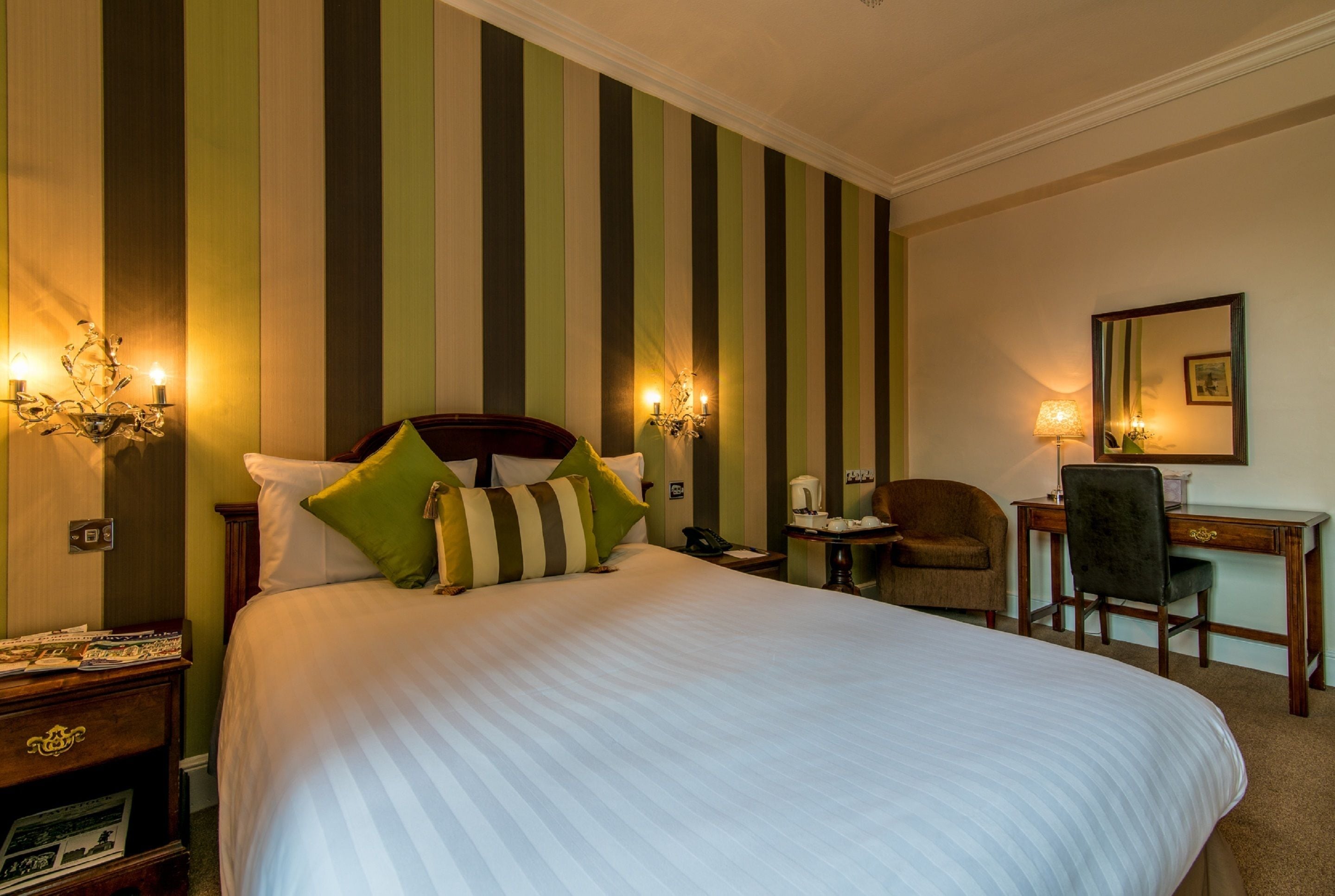 A hotel room with a large bed, striped wallpaper, a desk with a chair, and wall sconces.