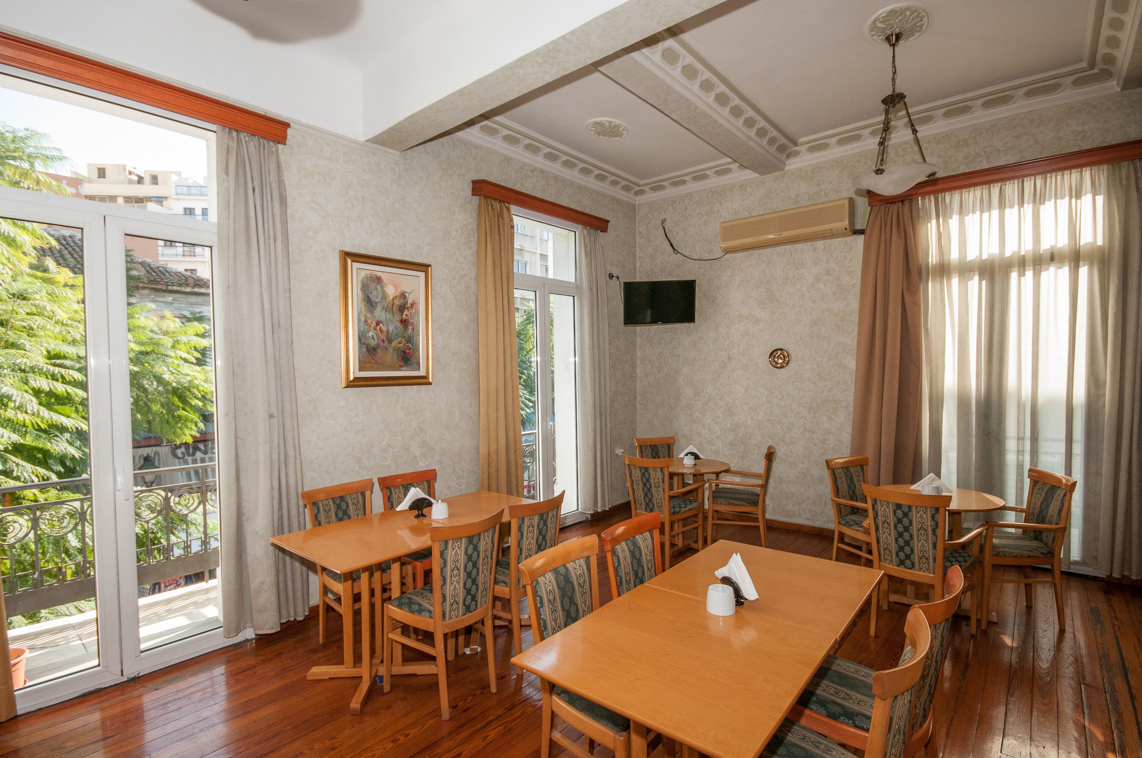 Uma sala de jantar com mesas e cadeiras de madeira, um quadro na parede, uma televisão e uma varanda com vista para árvores.