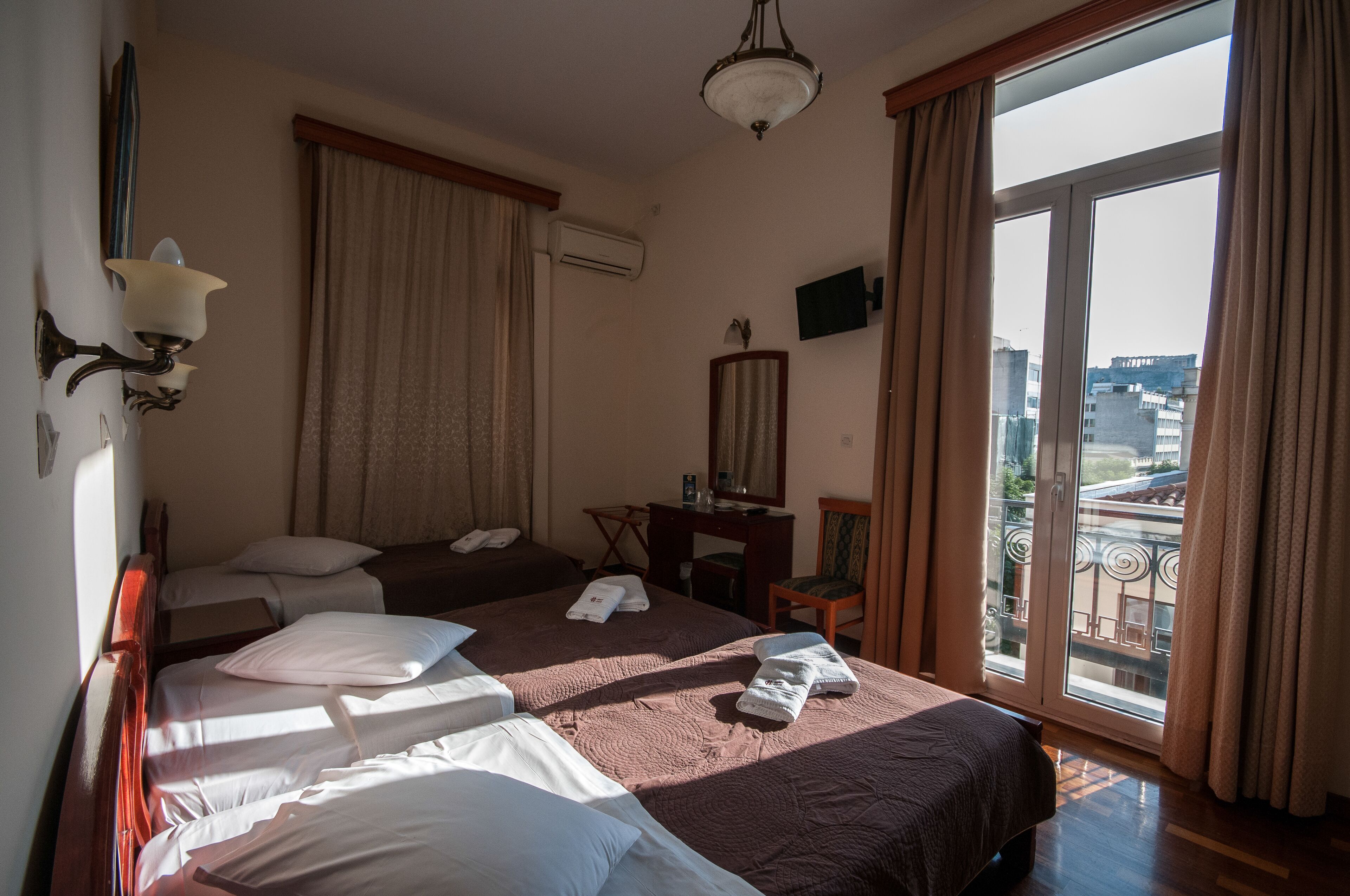 A hotel room with two beds, a desk, a chair, a TV, and a large window with a view of buildings.