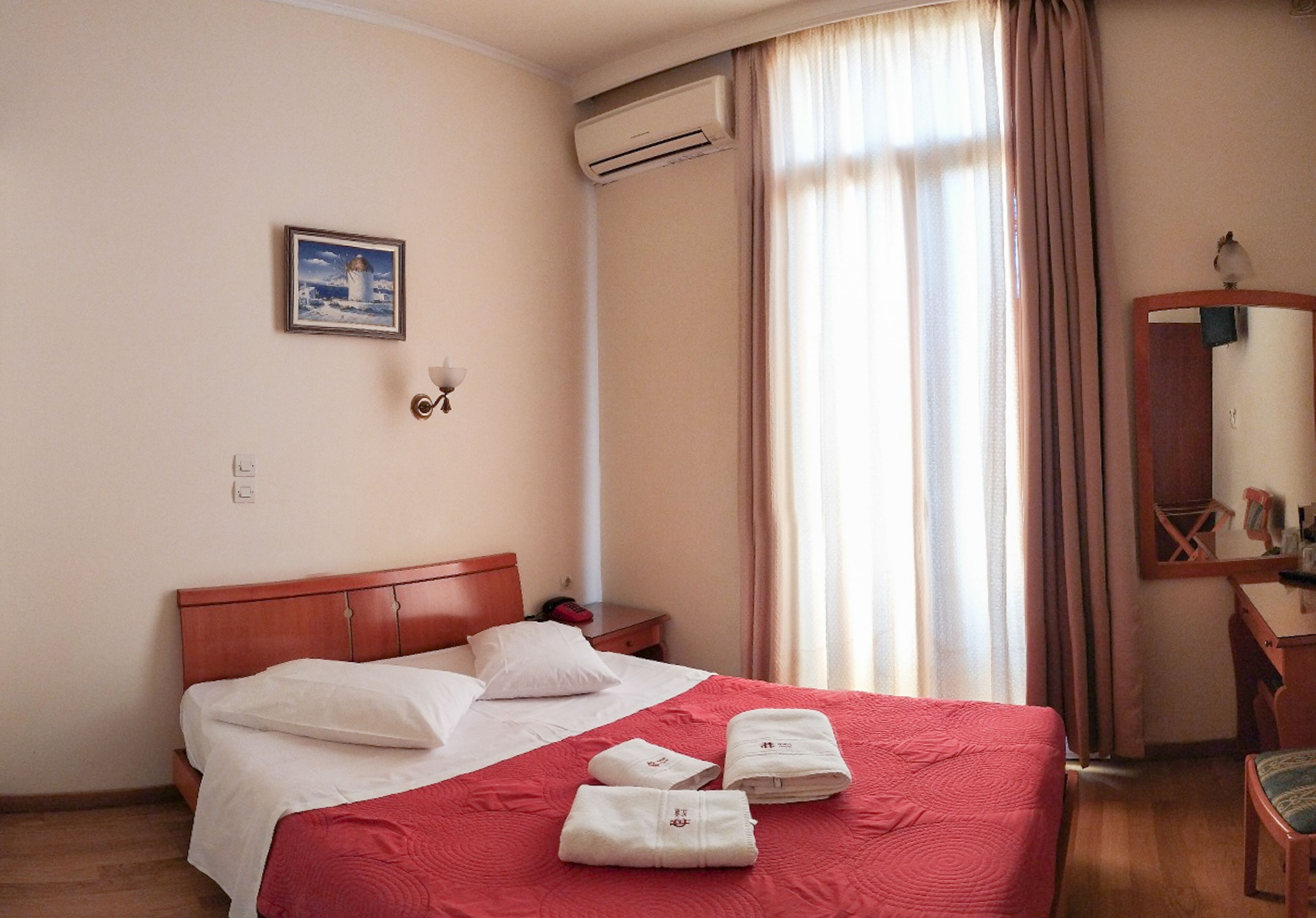 Habitación de hotel con cama, escritorio, silla, aire acondicionado, ventana con cortinas e imagen colgada en la pared.