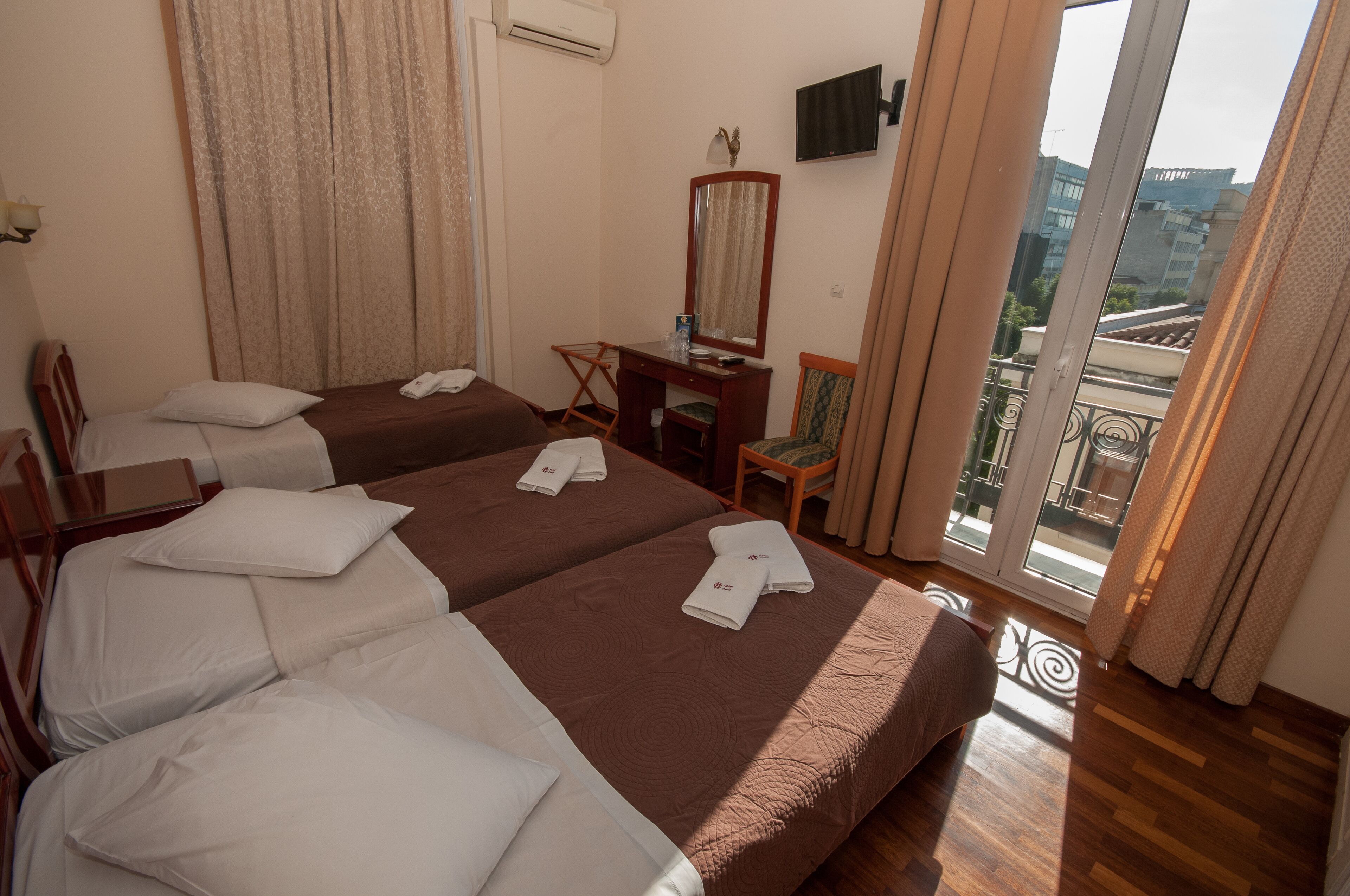 A hotel room with two beds, a desk, a chair, a TV, and a balcony with a view of buildings.