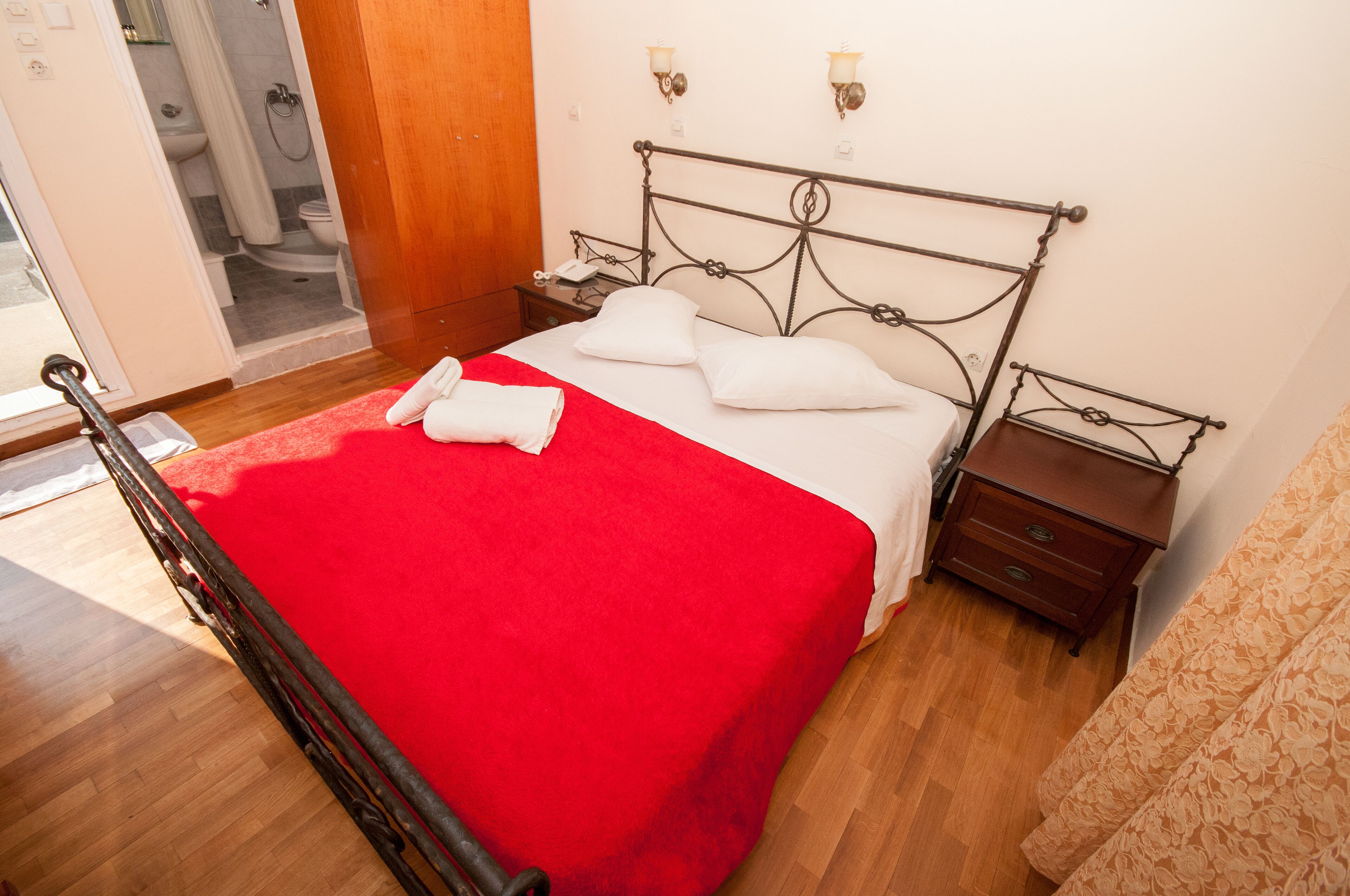 Un letto ben rifatto con coperta rossa, un comodino in legno con una lampada e un armadio in legno.
