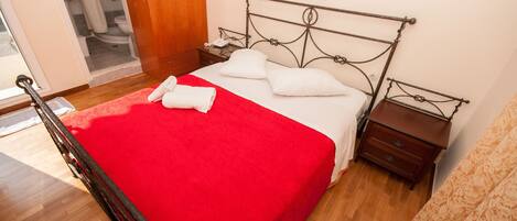 A neatly made bed with a red blanket, a wooden nightstand with a lamp, and a wooden wardrobe.