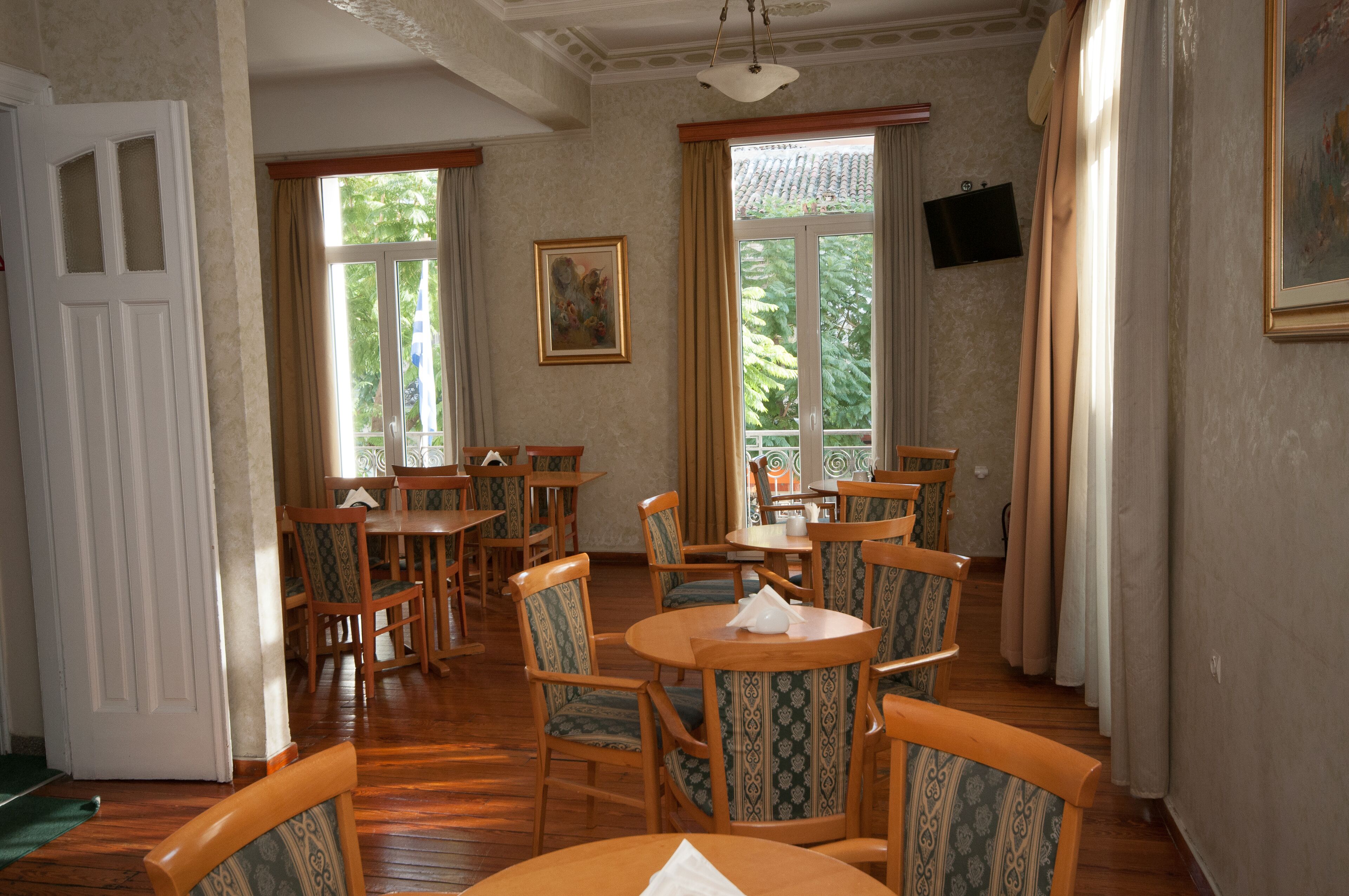 A dining area with wooden tables and chairs, large windows with curtains, and a painting on the wall.