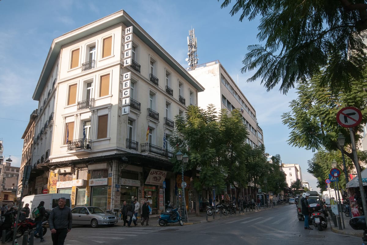 Hotel con balcón, cartel y vista a la calle con peatones y vehículos estacionados.
