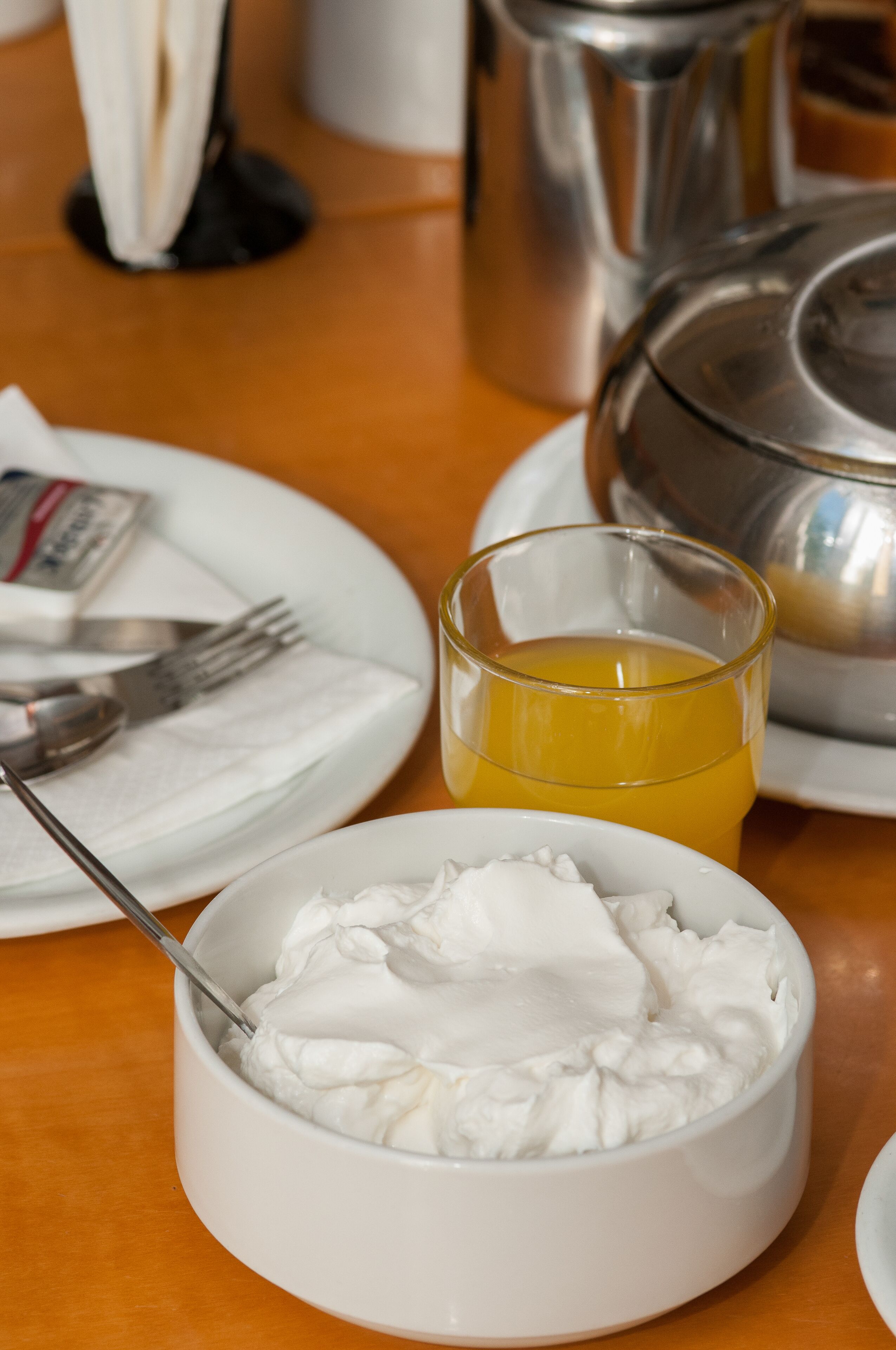 A dining table with a white bowl of whipped cream, a glass of orange juice, and a silver teapot.