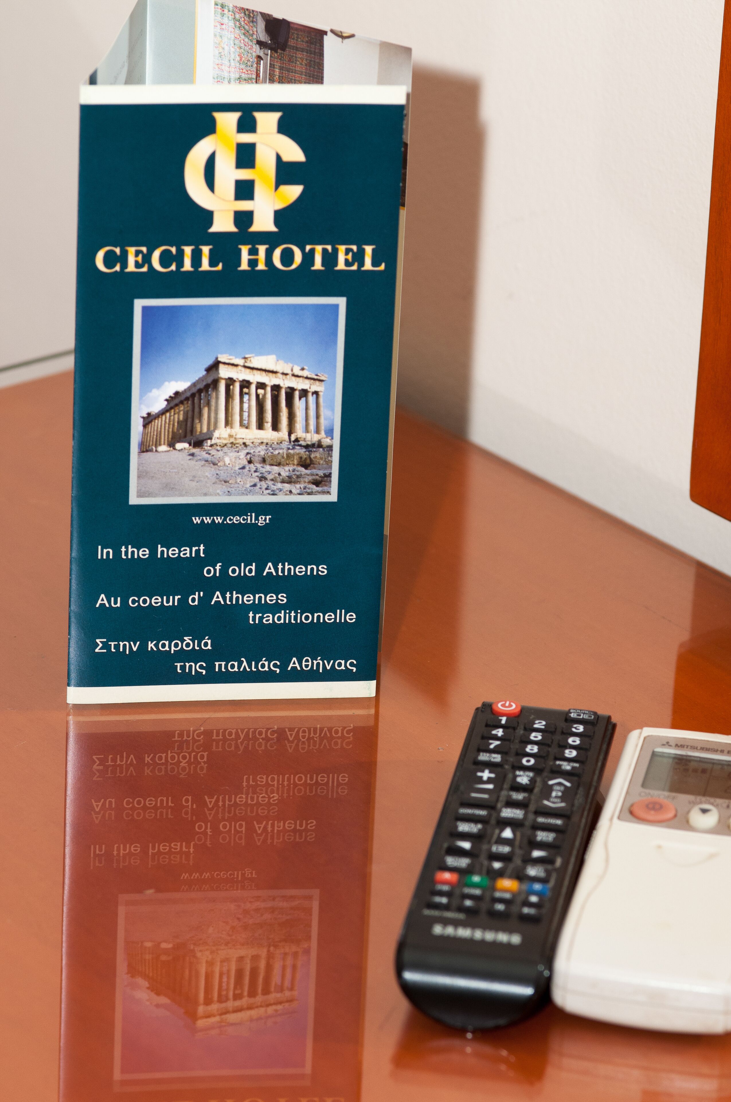A book titled 'CECIL HOTEL' with a photo of an ancient Greek building, a remote control, and a mobile phone on a wooden surface.