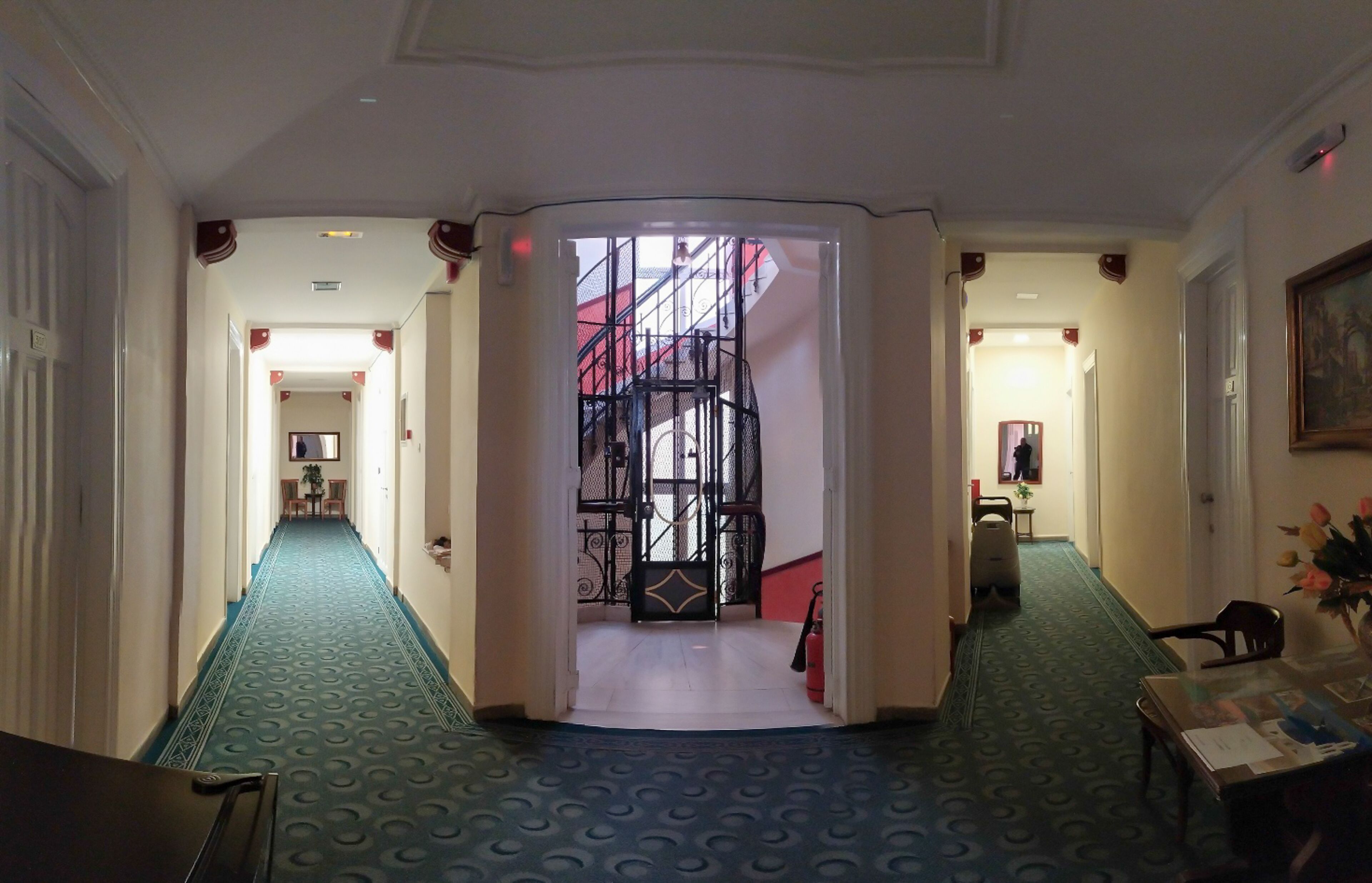 A hotel corridor with a blue carpet, a staircase with a red railing, and a painting on the wall.