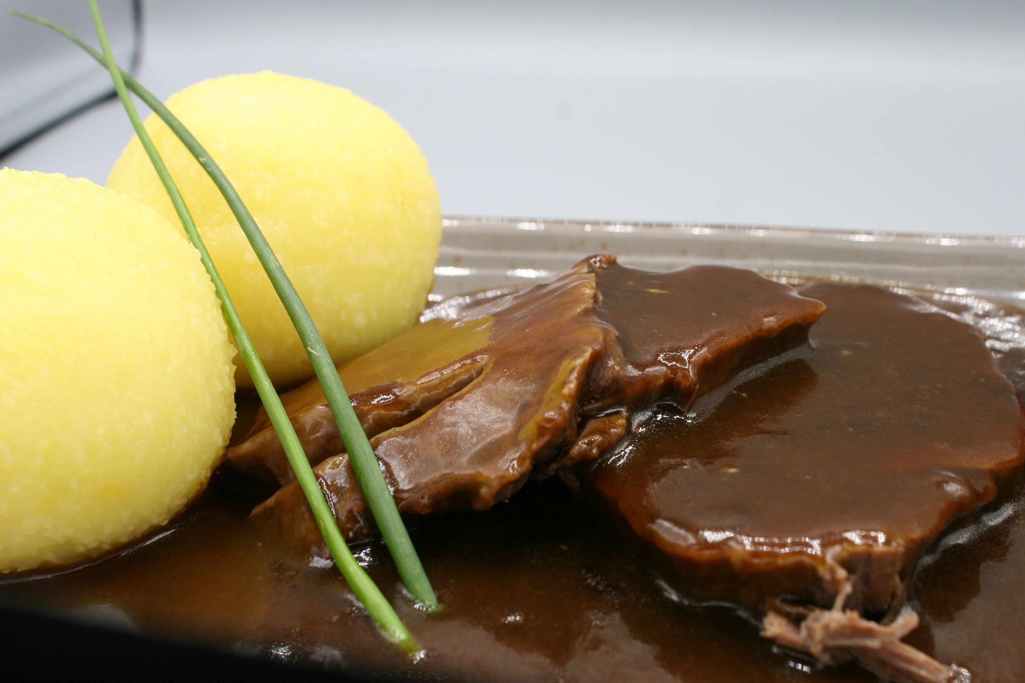 Un plato con un guiso de carne, posiblemente de res, cubierto con salsa marrón, acompañado de dos albóndigas amarillas y una guarnición verde.