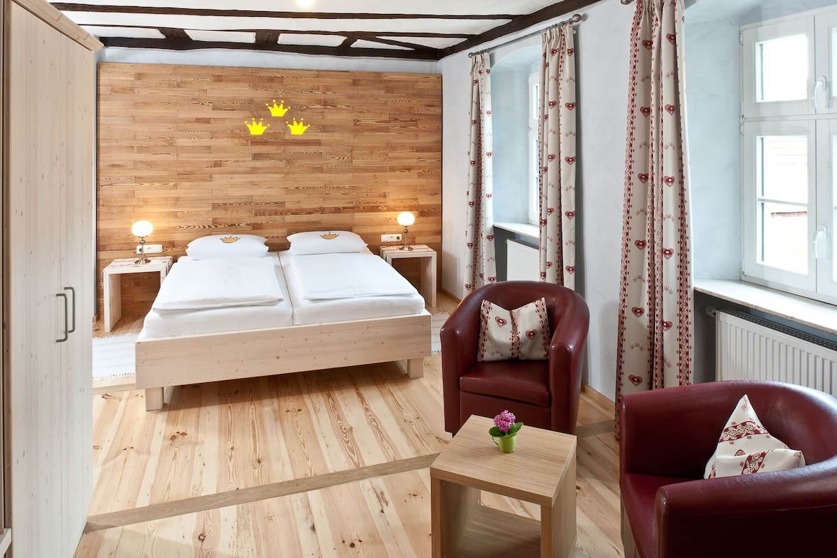 A bedroom with a wooden headboard, a double bed, two bedside lamps, a red leather armchair, a small wooden table with a flower, and a window with curtains.