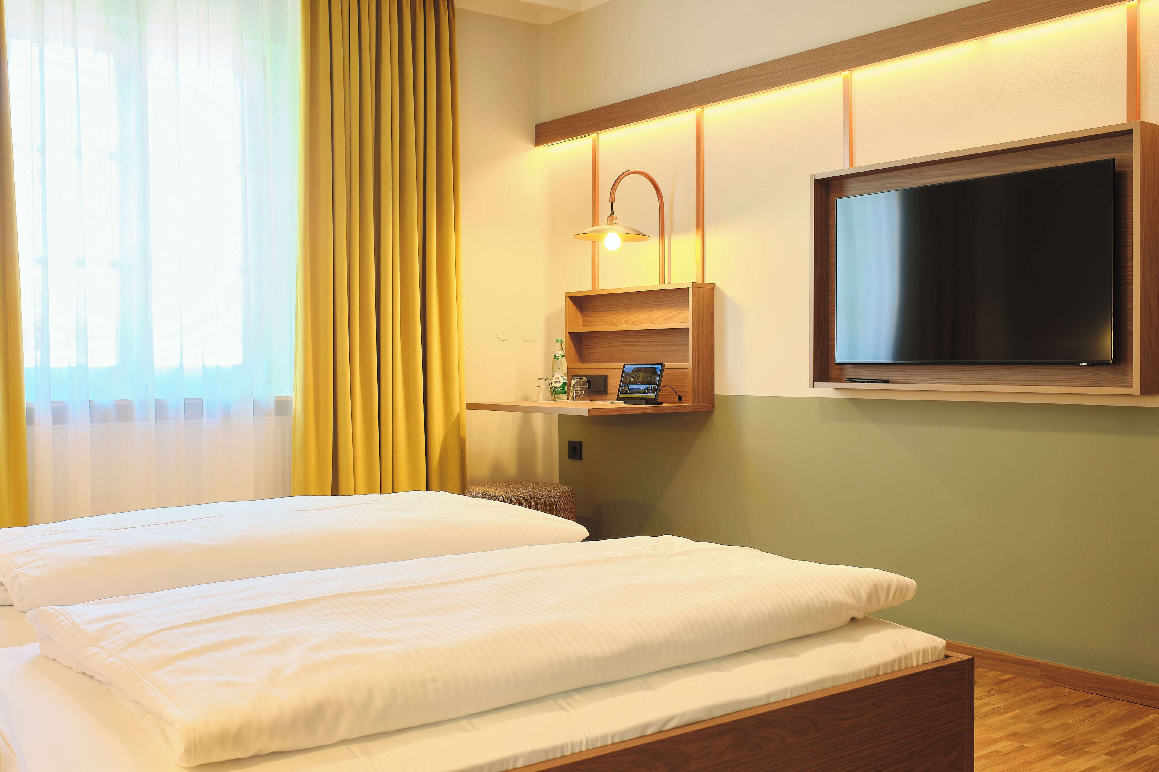 A hotel room with two beds, a TV mounted on the wall, and a bedside table with a lamp.