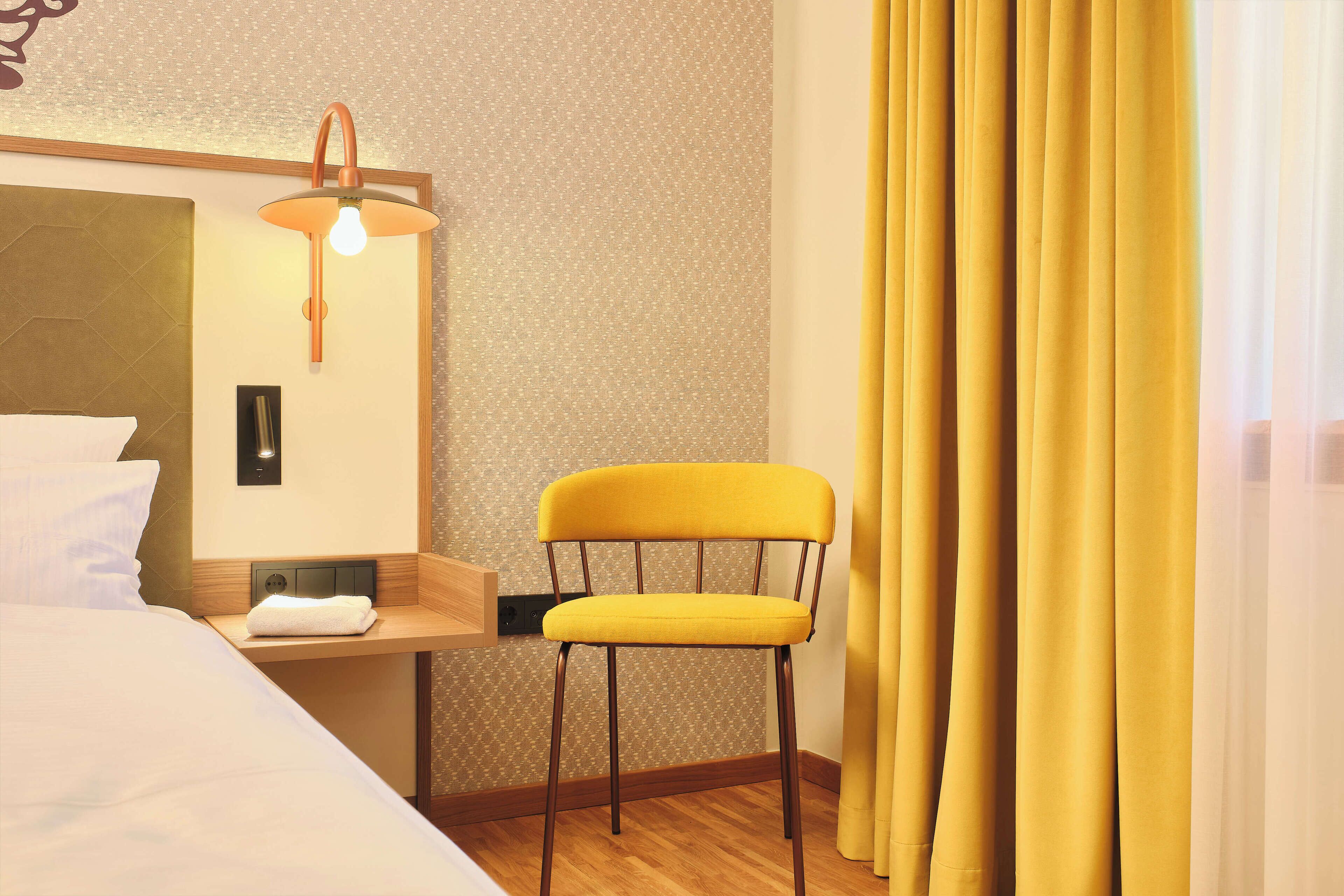A hotel room with a bed, a chair, a nightstand, a lamp, and curtains.