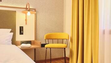 A hotel room with a bed, a chair, a nightstand, a lamp, and curtains.