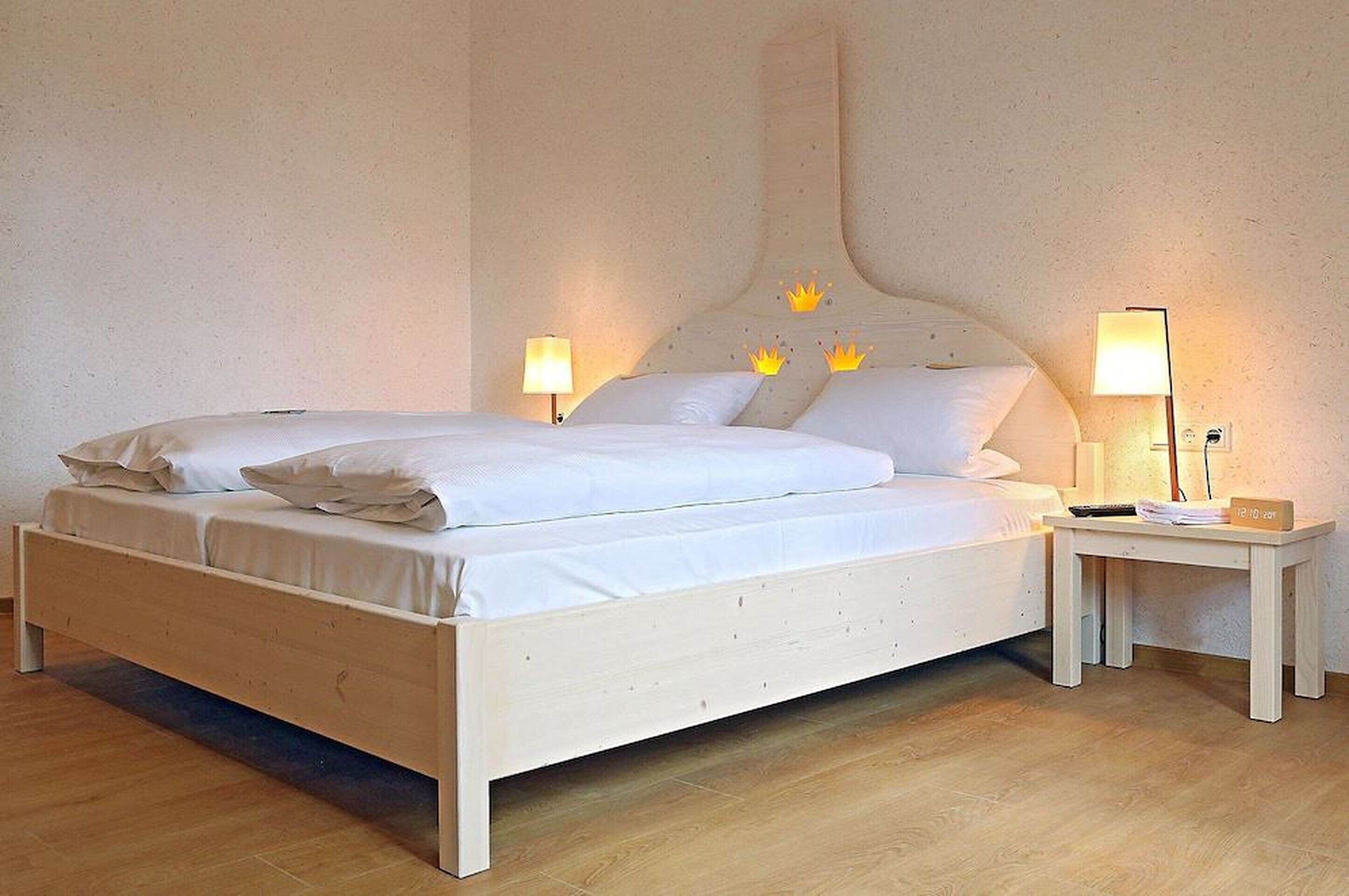 Een tweepersoonsbed met witte lakens, twee nachtkastjes met lampen, en een klein nachtkastje met een klok.