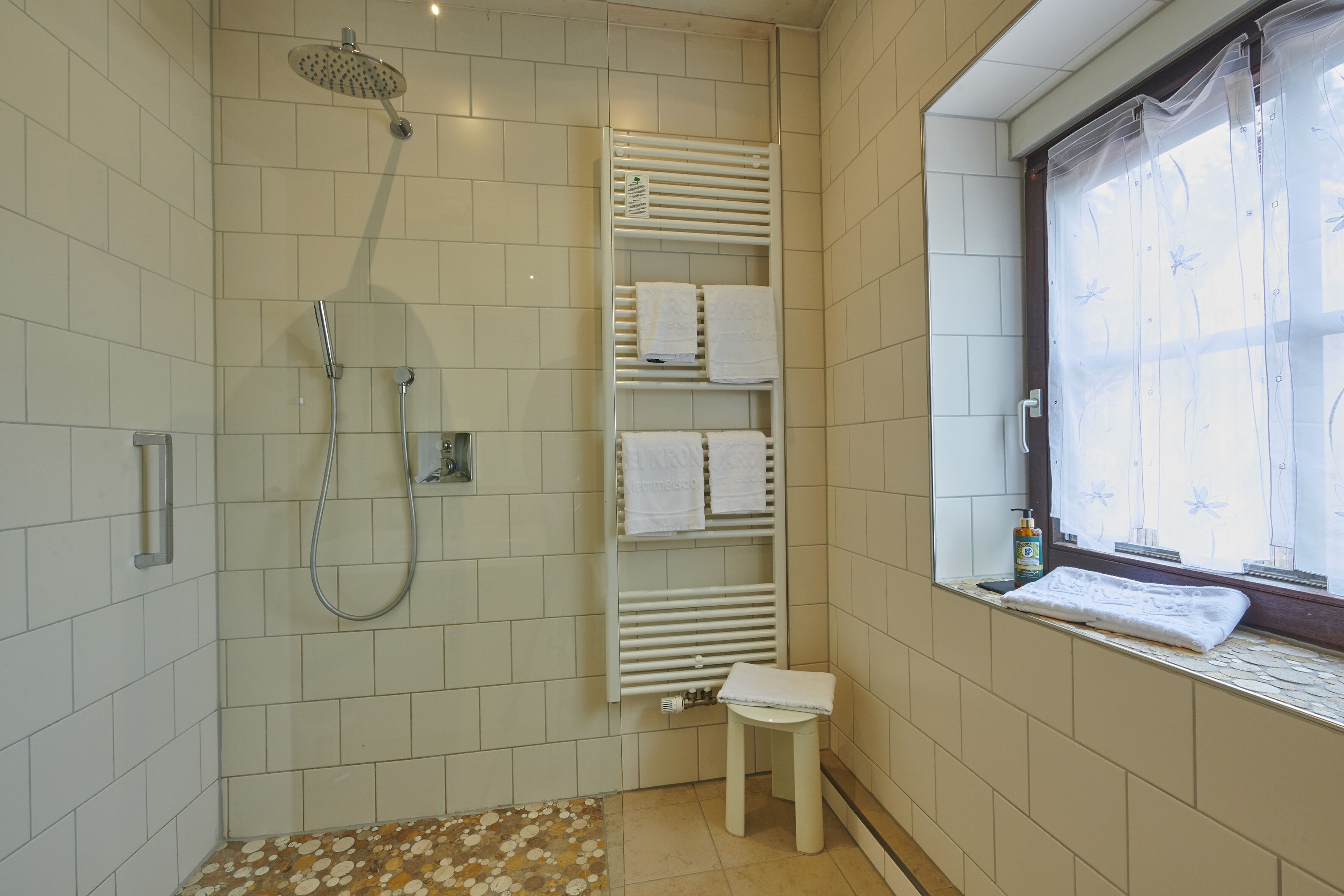 A bathroom with a shower, a towel rack, a small stool, and a window with a view.