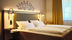 Hotellihuone, jossa on suuri sänky, kaksi yöpöytävalaisinta, yöpöytä, tuoli ja seinässä kruunukuviolla koristeltu elementti.