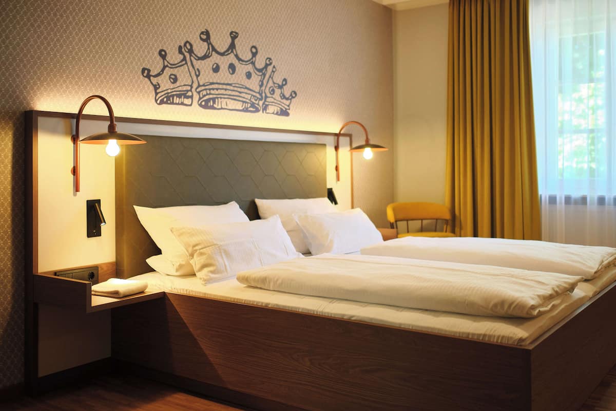A hotel room with a large bed, two bedside lamps, a nightstand, a chair, and a crown design on the wall.