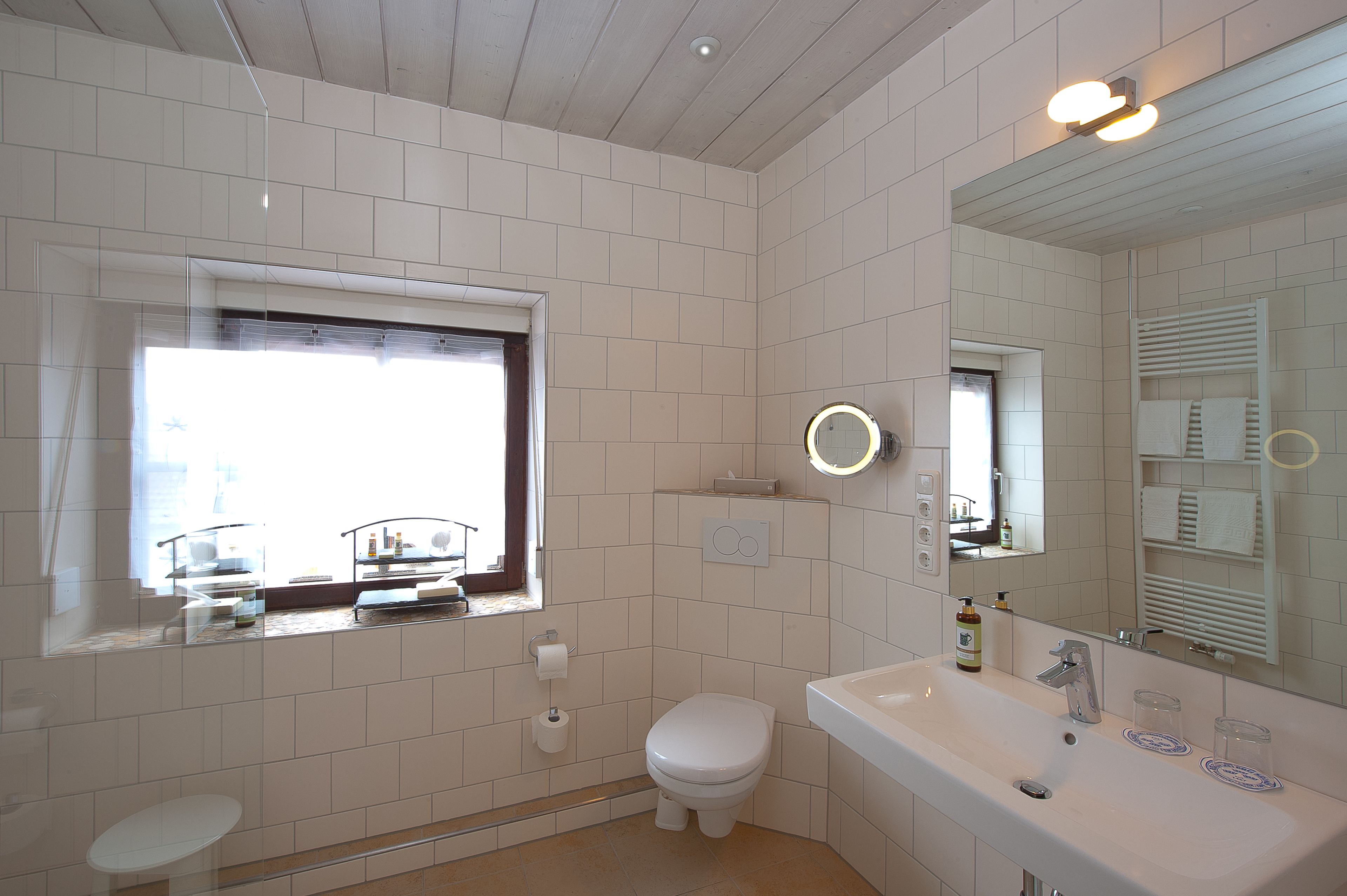 A bathroom with white tiles, a toilet, a sink with a mirror, and a window.