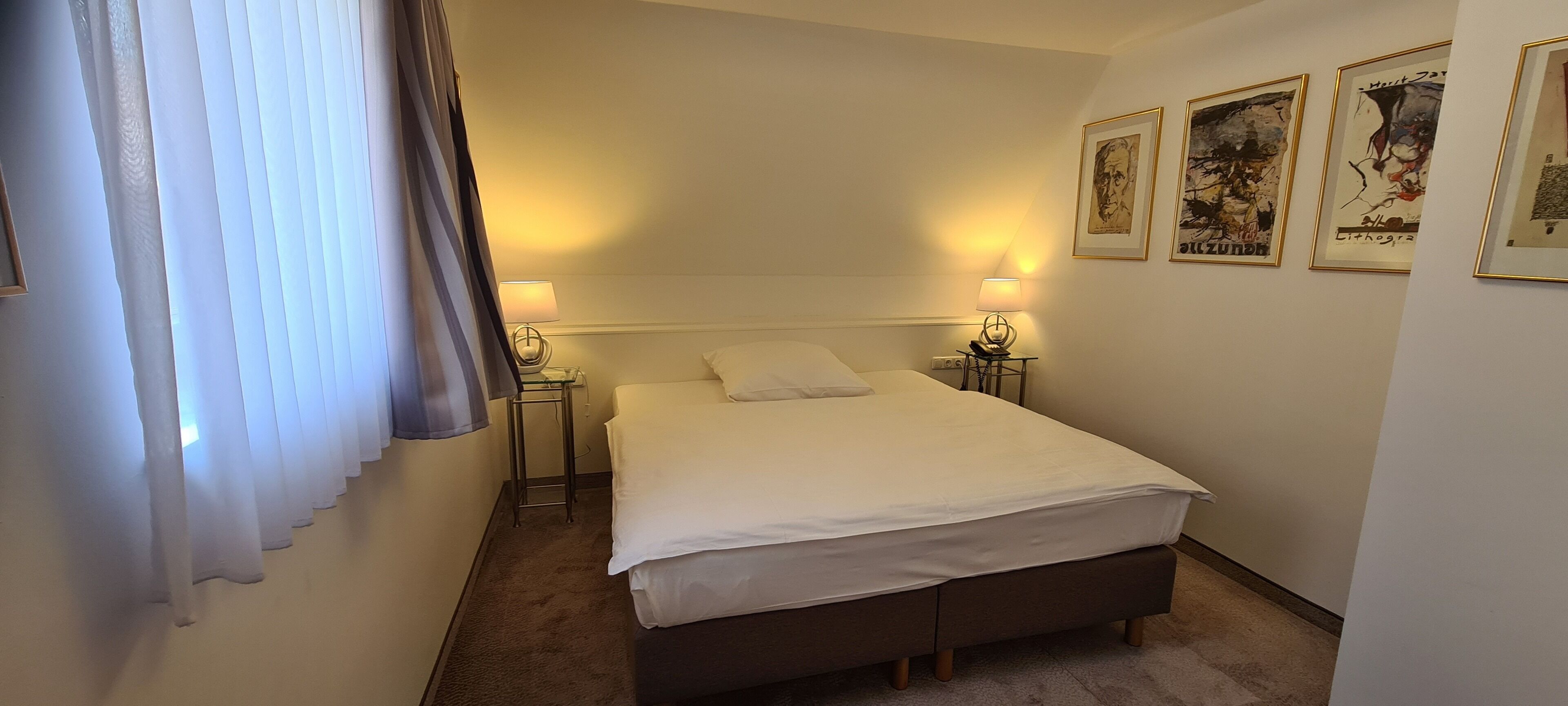 A neatly made bed in a hotel room with two bedside tables, each with a lamp. There are framed artworks on the wall.