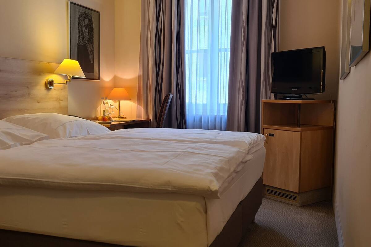 A hotel room with a double bed, two bedside lamps, a TV on a wooden stand, and a window with curtains.