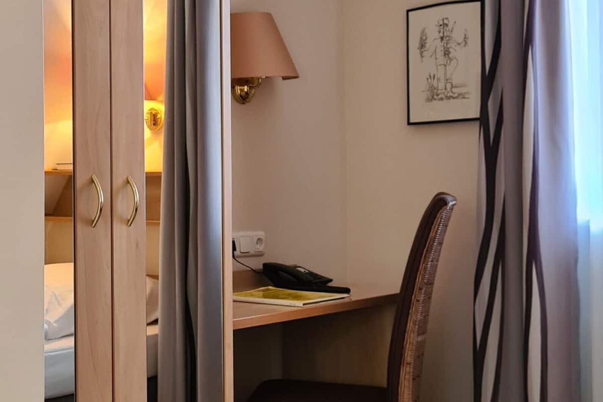 A hotel room with a wooden wardrobe, a desk with a chair, a lamp, and a framed picture on the wall.
