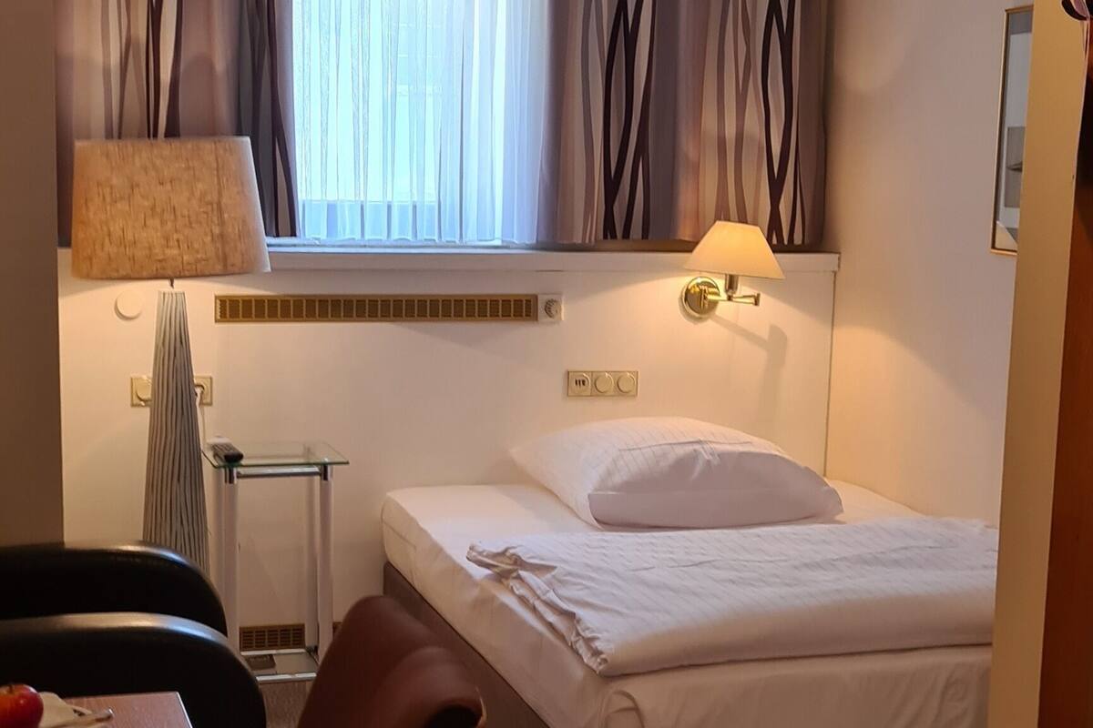A hotel room with a bed, a desk, a chair, a lamp, and a window with curtains.