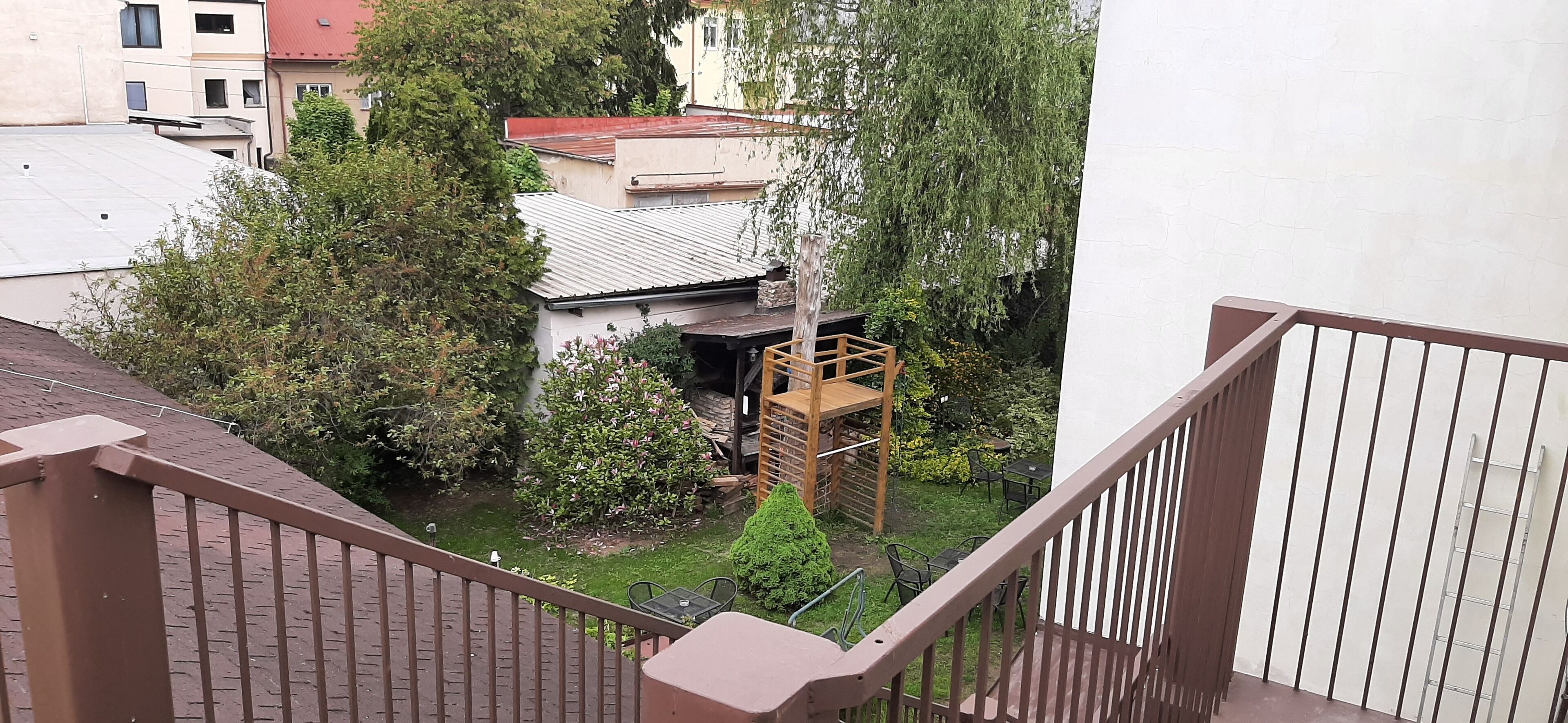A balcony with a view of a garden, a wooden deck, and a building with a red roof.