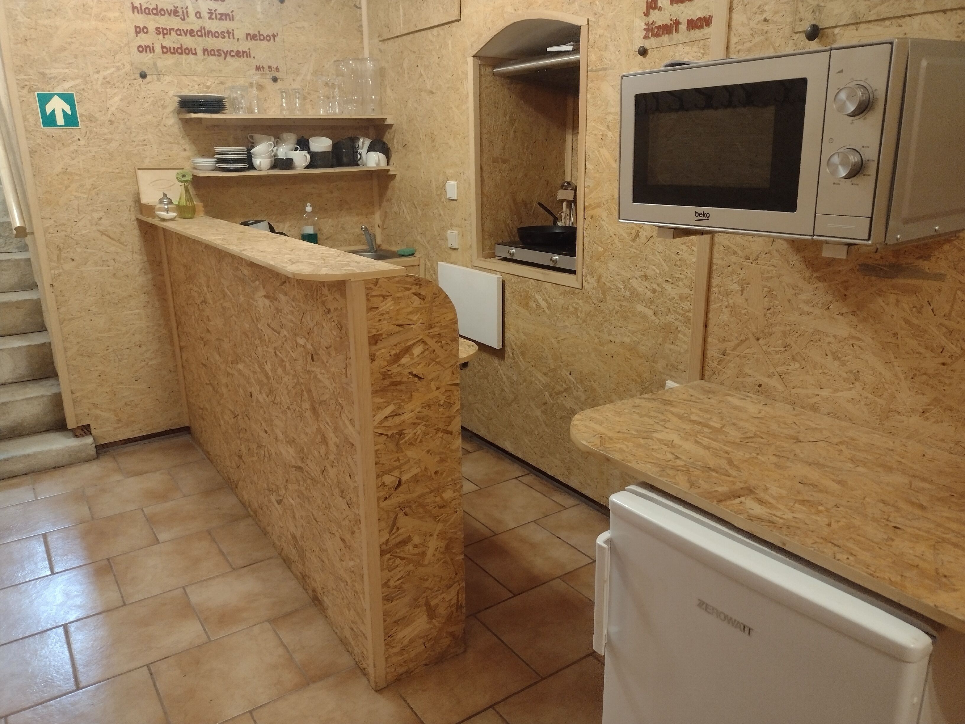 A kitchen with a wooden countertop, a microwave, and a refrigerator. There is a sink with a faucet and a dish rack with plates and cups. A sign with text is visible on the wall.