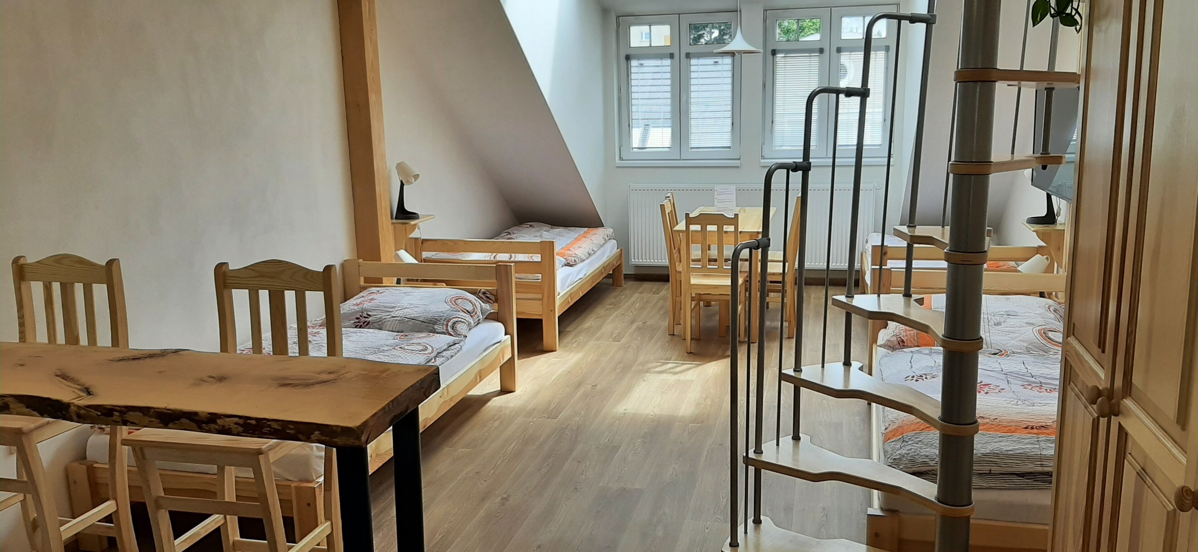 A dormitory room with bunk beds, a wooden table, chairs, a desk, and a ladder.