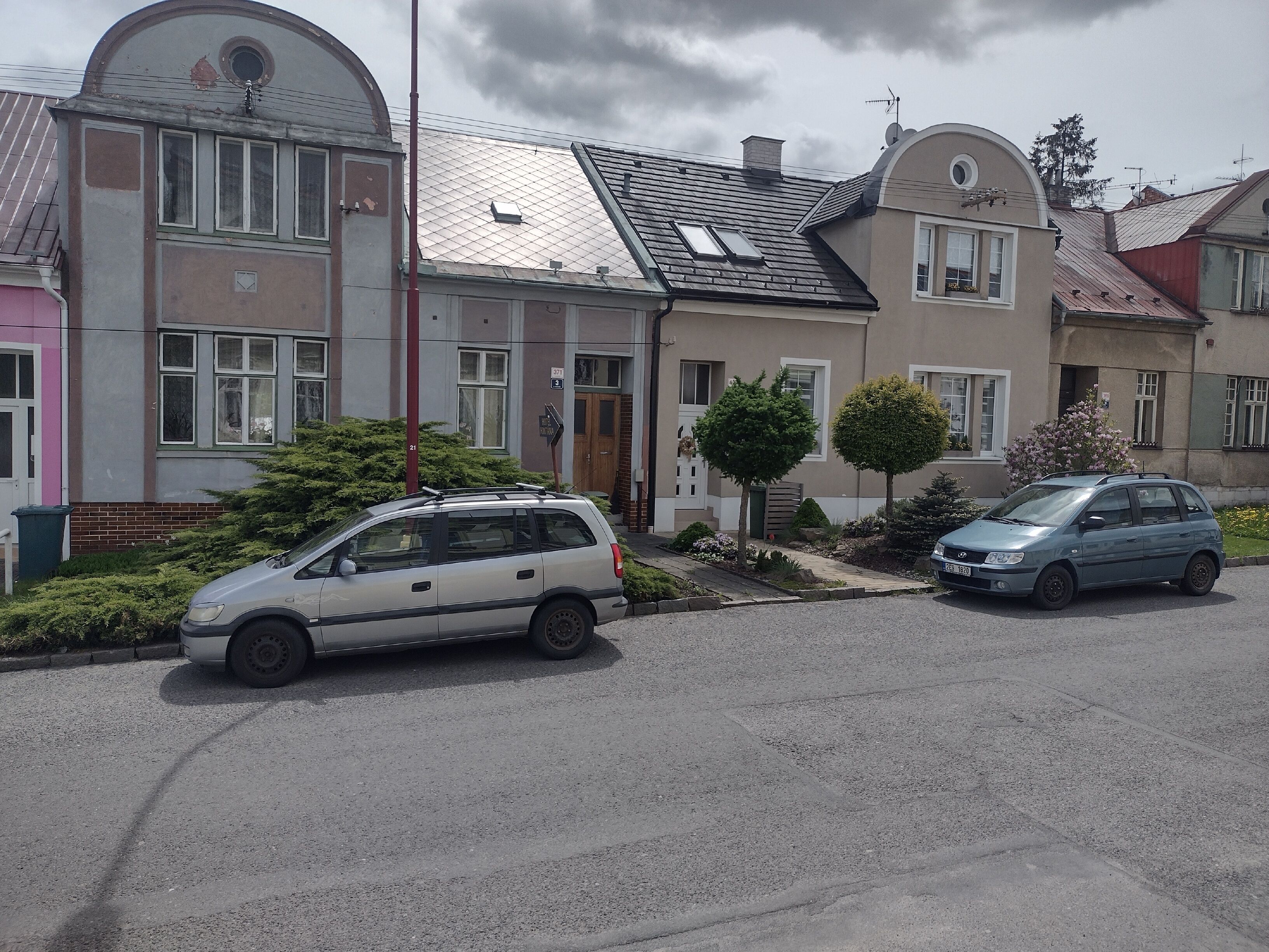 A residential area with two parked cars, a house with a sloped roof, and a small garden with trimmed bushes and a tree.