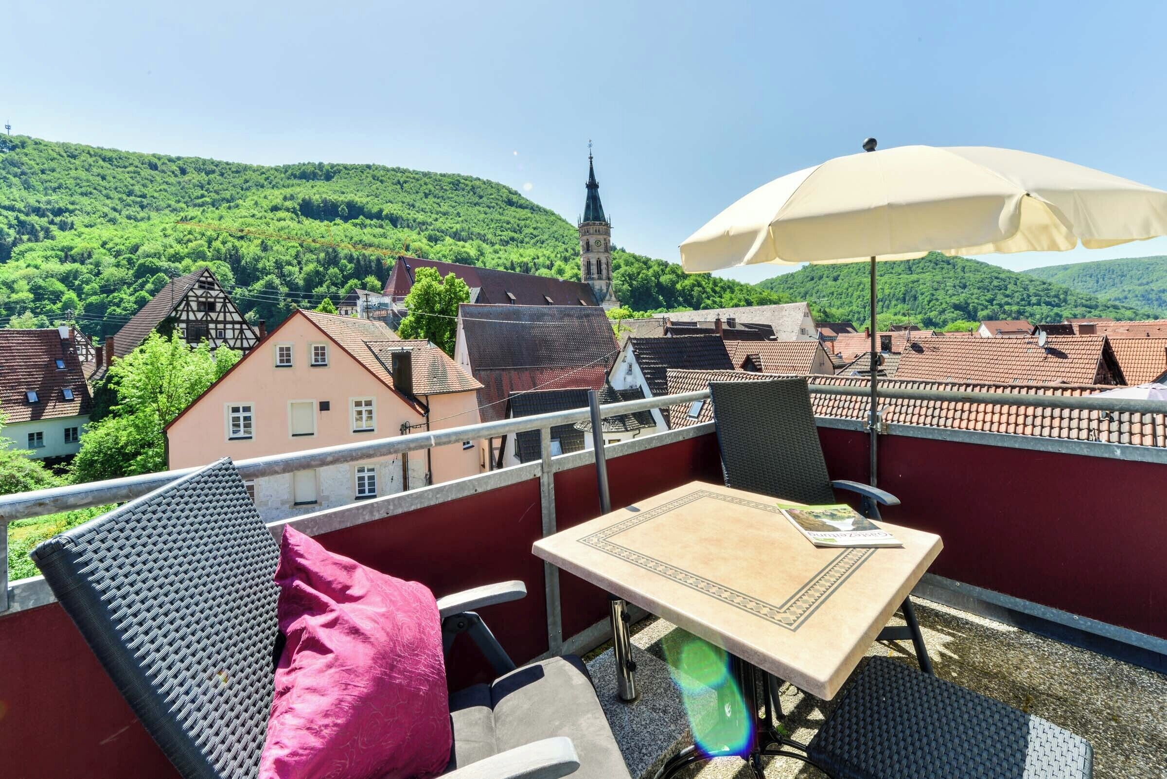 A balcony with a table, chairs, and an umbrella. The balcony overlooks a town with traditional buildings and a church tower.