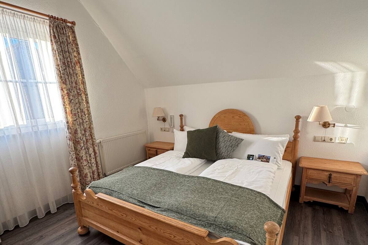 Una cama de madera con una colcha verde, ropa de cama blanca y un cabecero de madera. Hay dos mesitas de noche con lámparas, una ventana con cortinas y un candelabro.