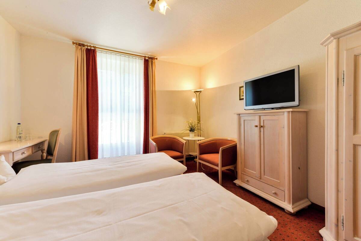 Habitación de hotel con dos camas, un escritorio, una silla, un televisor sobre un mueble y una ventana con cortinas.